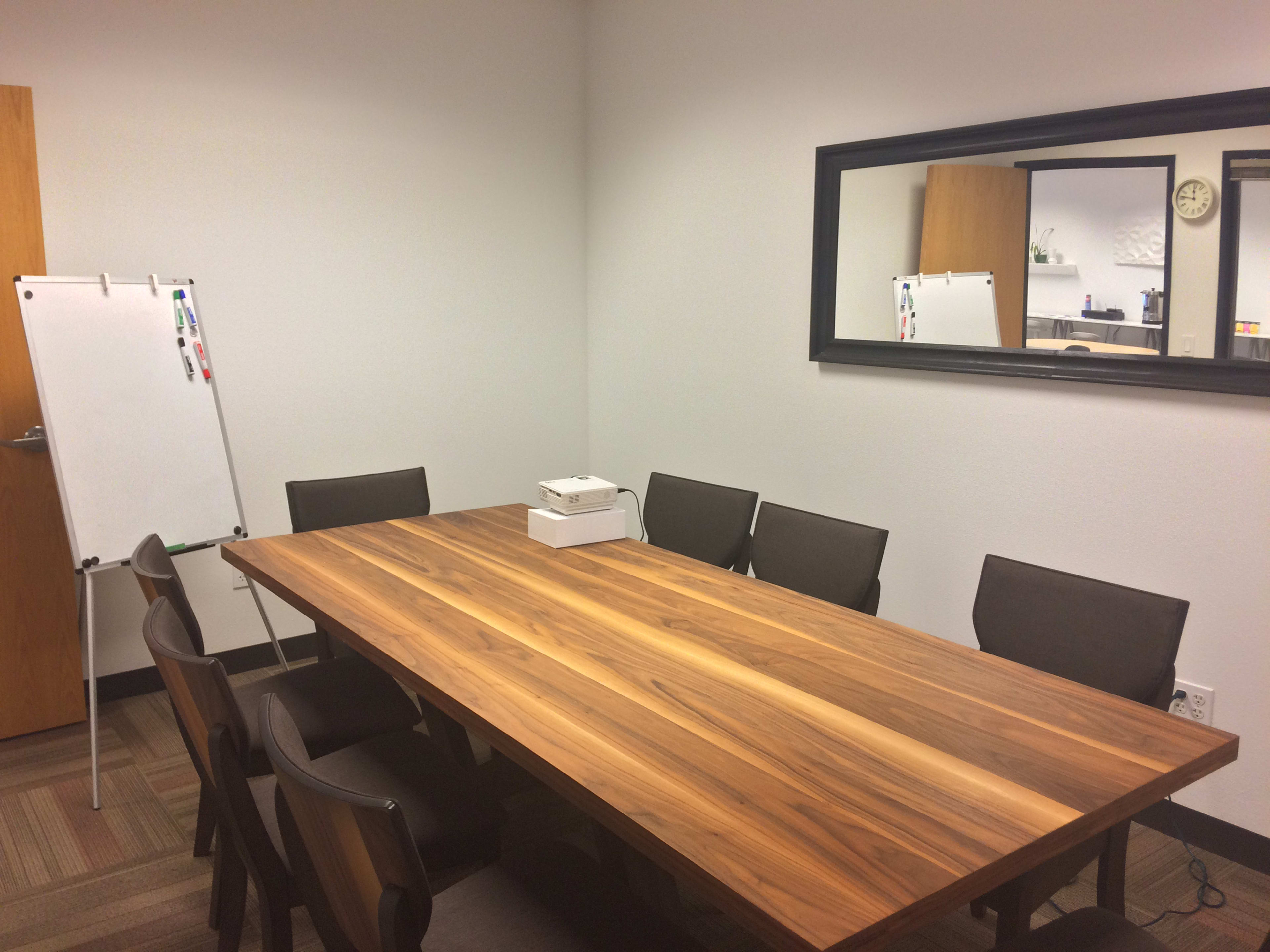A conference room features a long wooden table surrounded by eight chairs, with a whiteboard and a mirror on the walls.