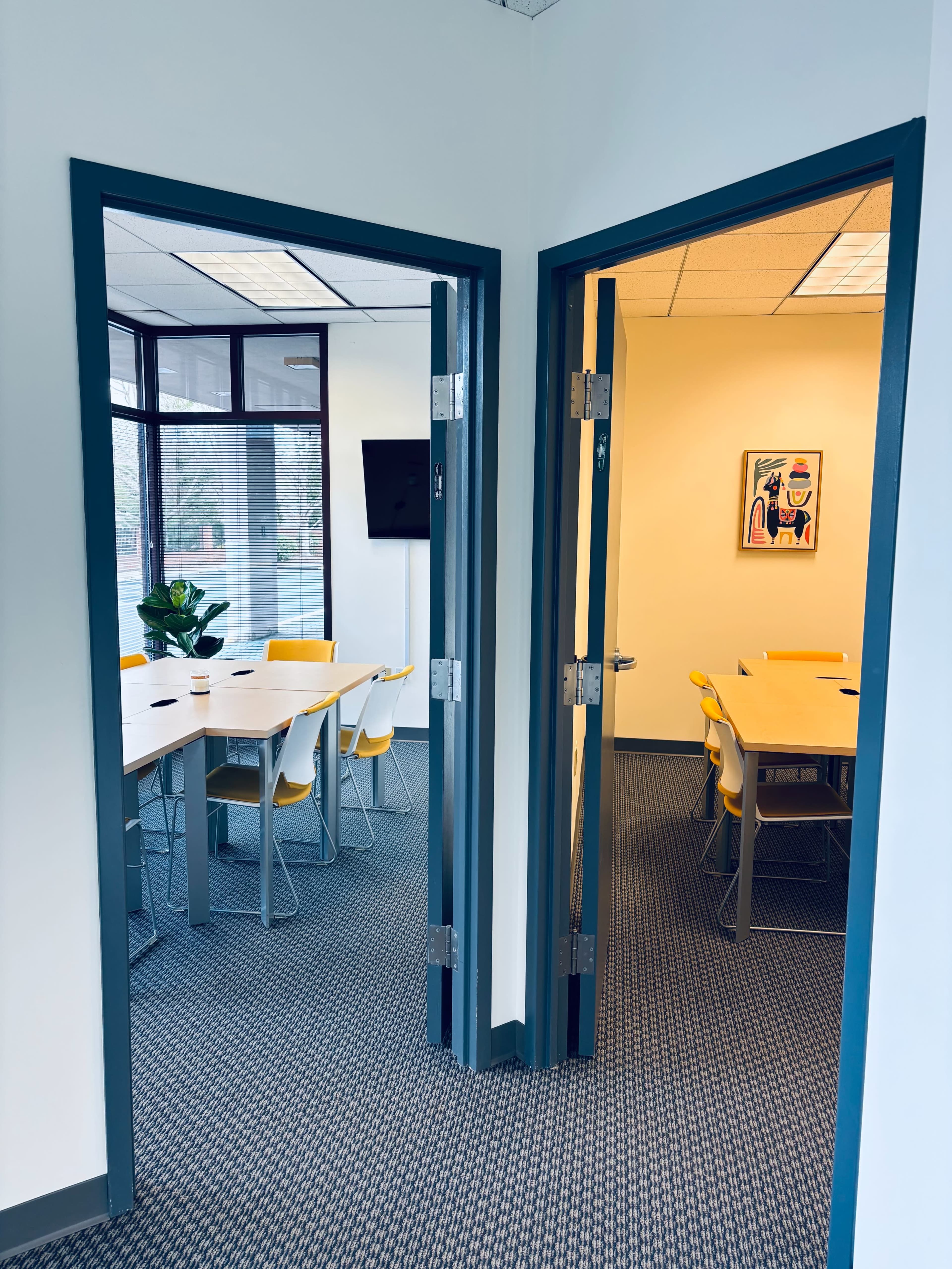 The image shows a corridor with two open doors leading to conference rooms, one featuring a long table and yellow chairs, and the other a more traditional seating arrangement.