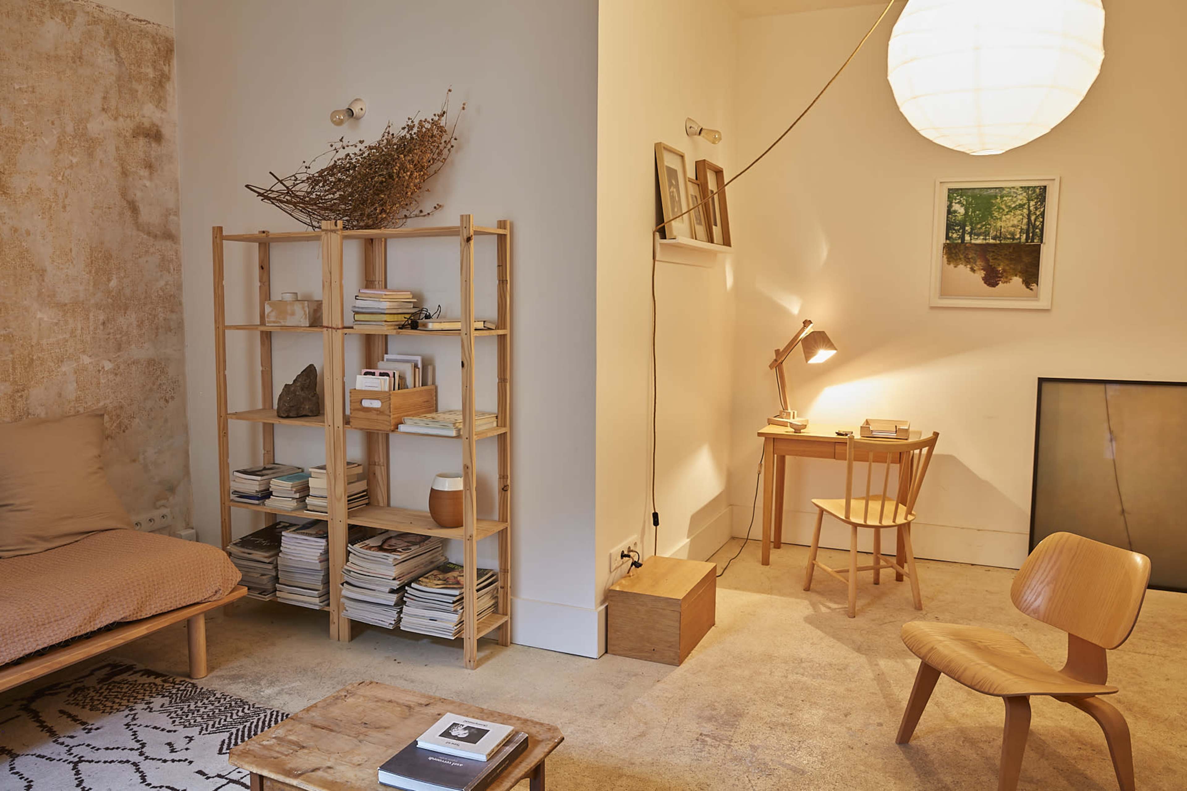 The image displays a softly lit room featuring a wooden bookshelf filled with books, a cozy bed, a small desk with a chair, and a round lamp hanging softly from the ceiling.