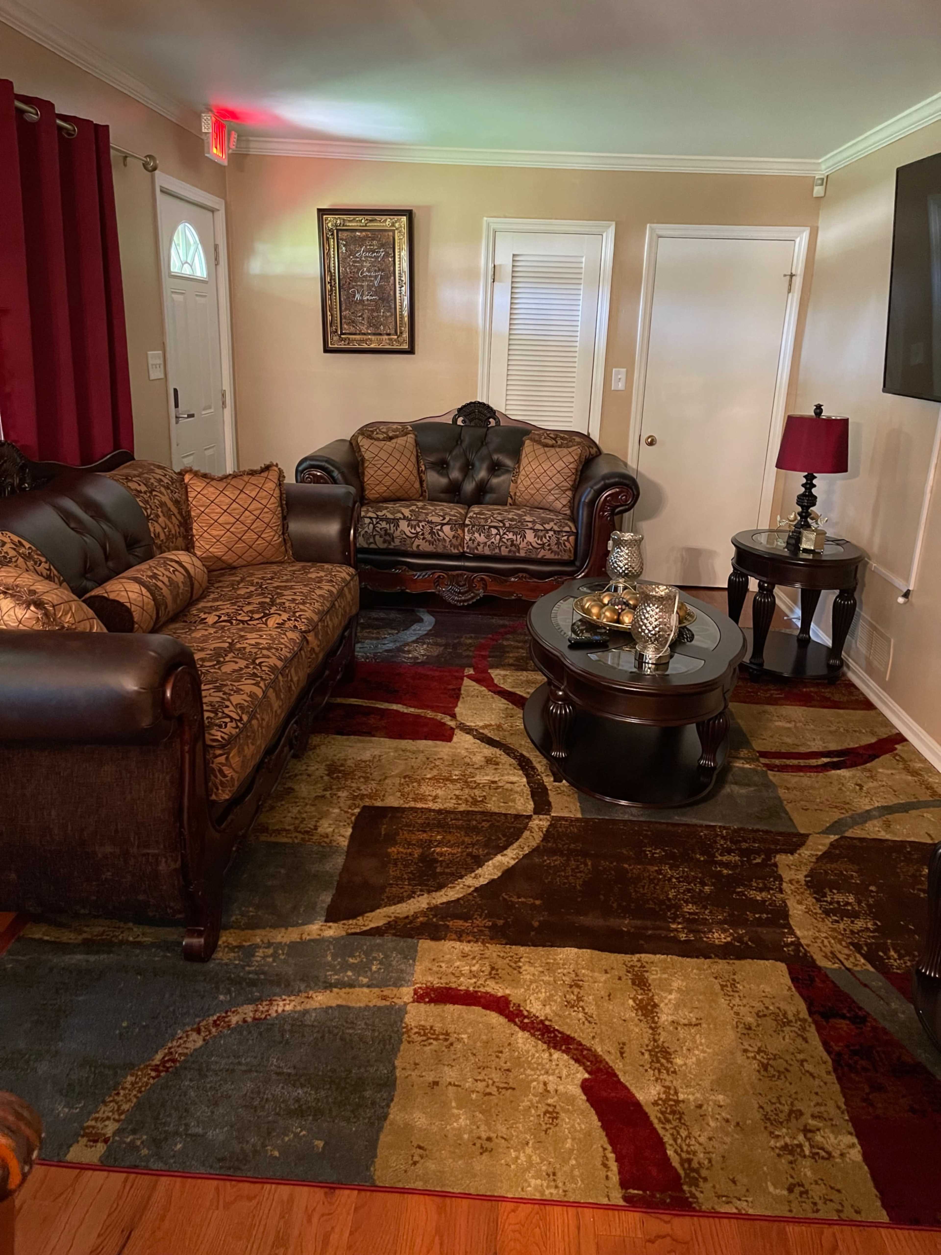 A living room with a brown leather sofa and loveseat, a round glass coffee table, and a colorful rug with circular patterns.