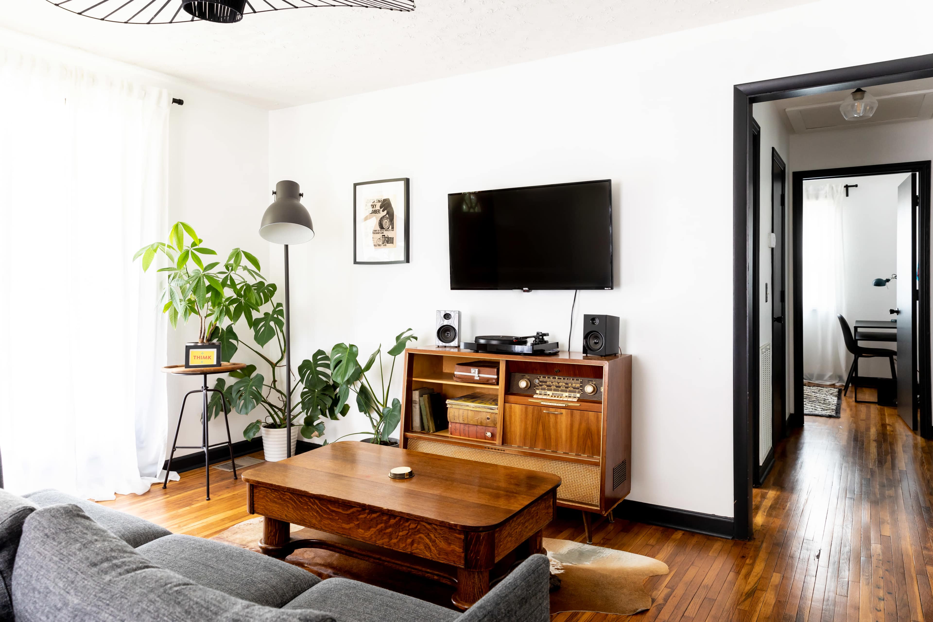 A living room features a grey sofa, wooden coffee table, a vintage-style media console with a television, speakers, and plants, against white walls and hardwood flooring.