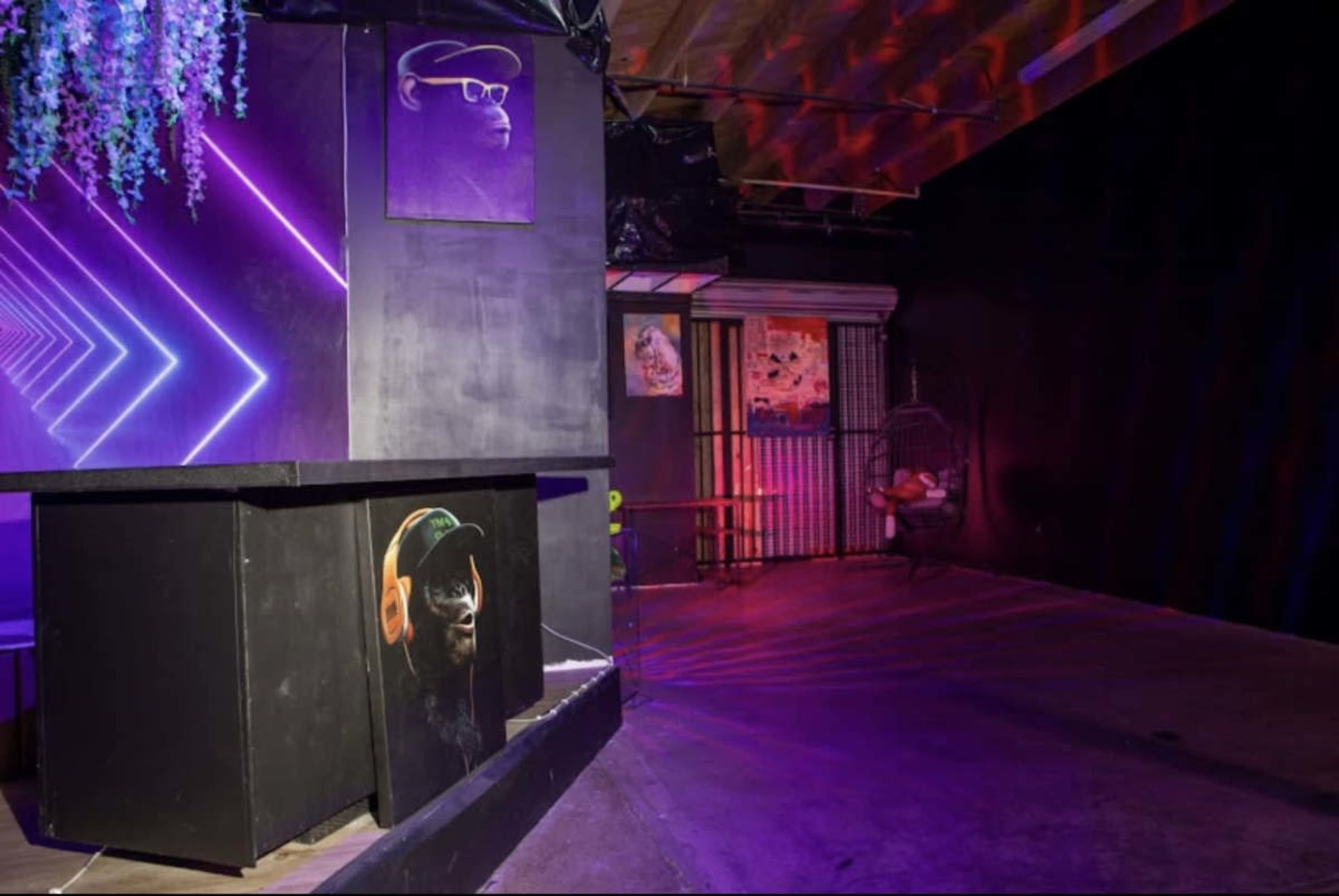 The scene features a dimly lit room with purple neon lighting, showcasing a bar area with a mural of a monkey wearing headphones on the front and a hanging chair in the corner.