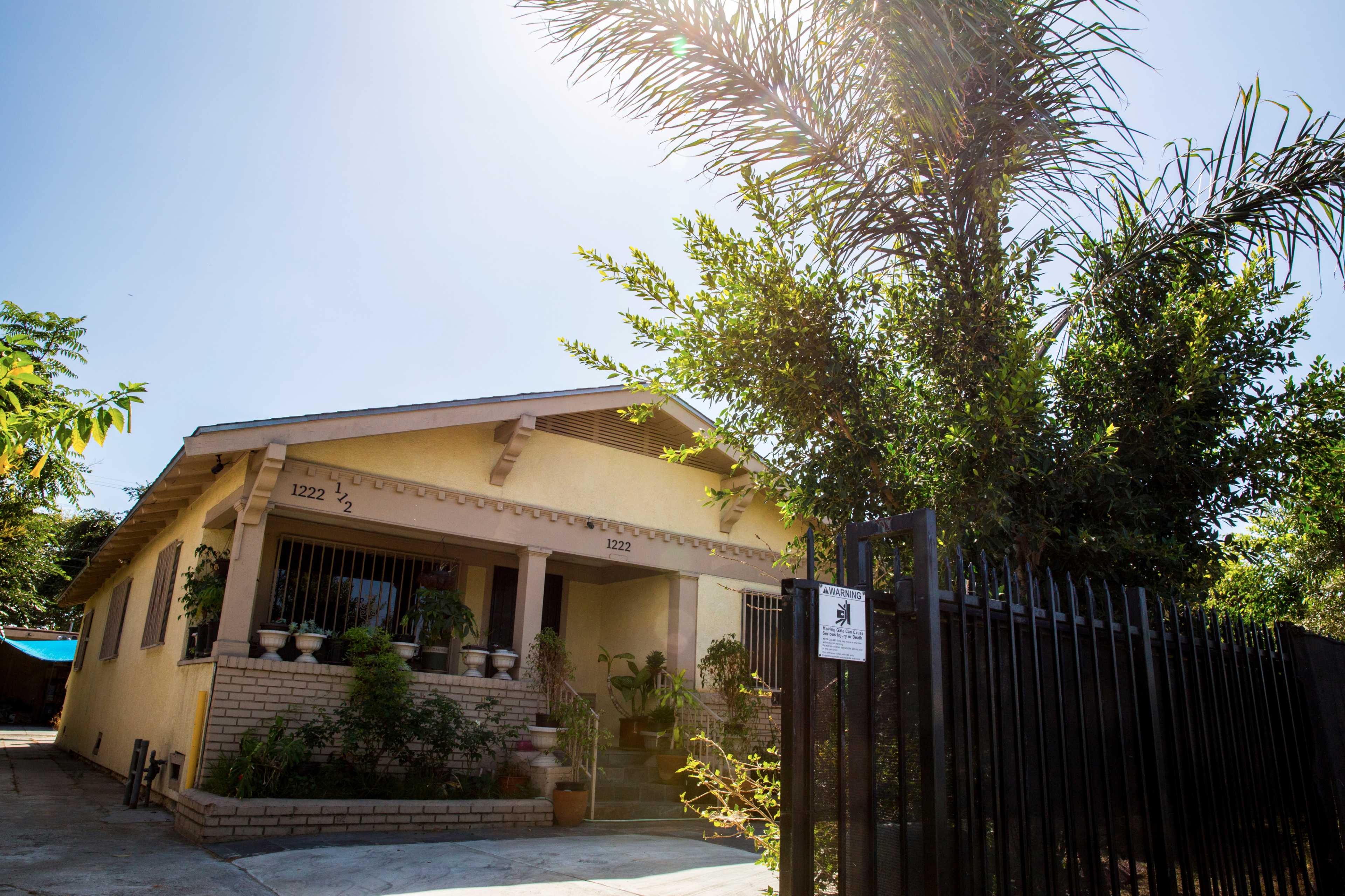 A single-story house with a fenced yard and potted plants on the porch is set against a clear sky.