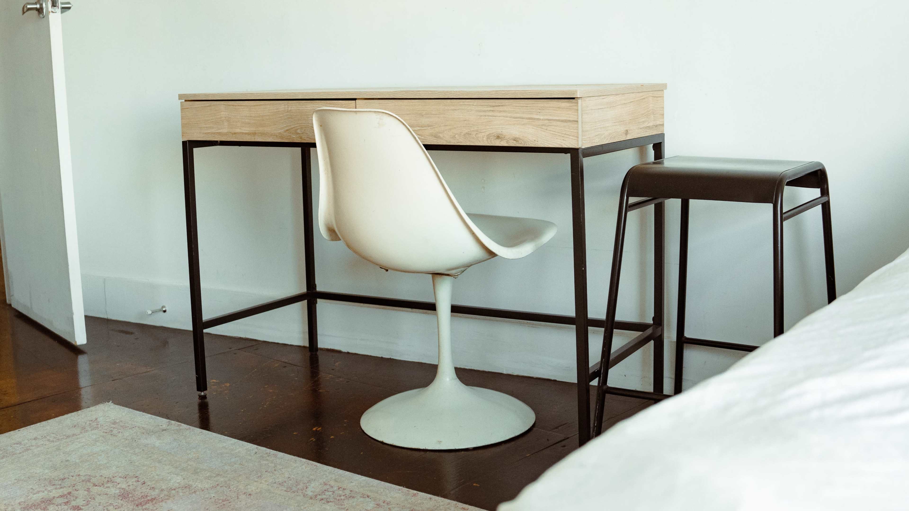 A wooden desk with a drawer is paired with a white, pedestal-style chair and a black stool in a minimalist room.