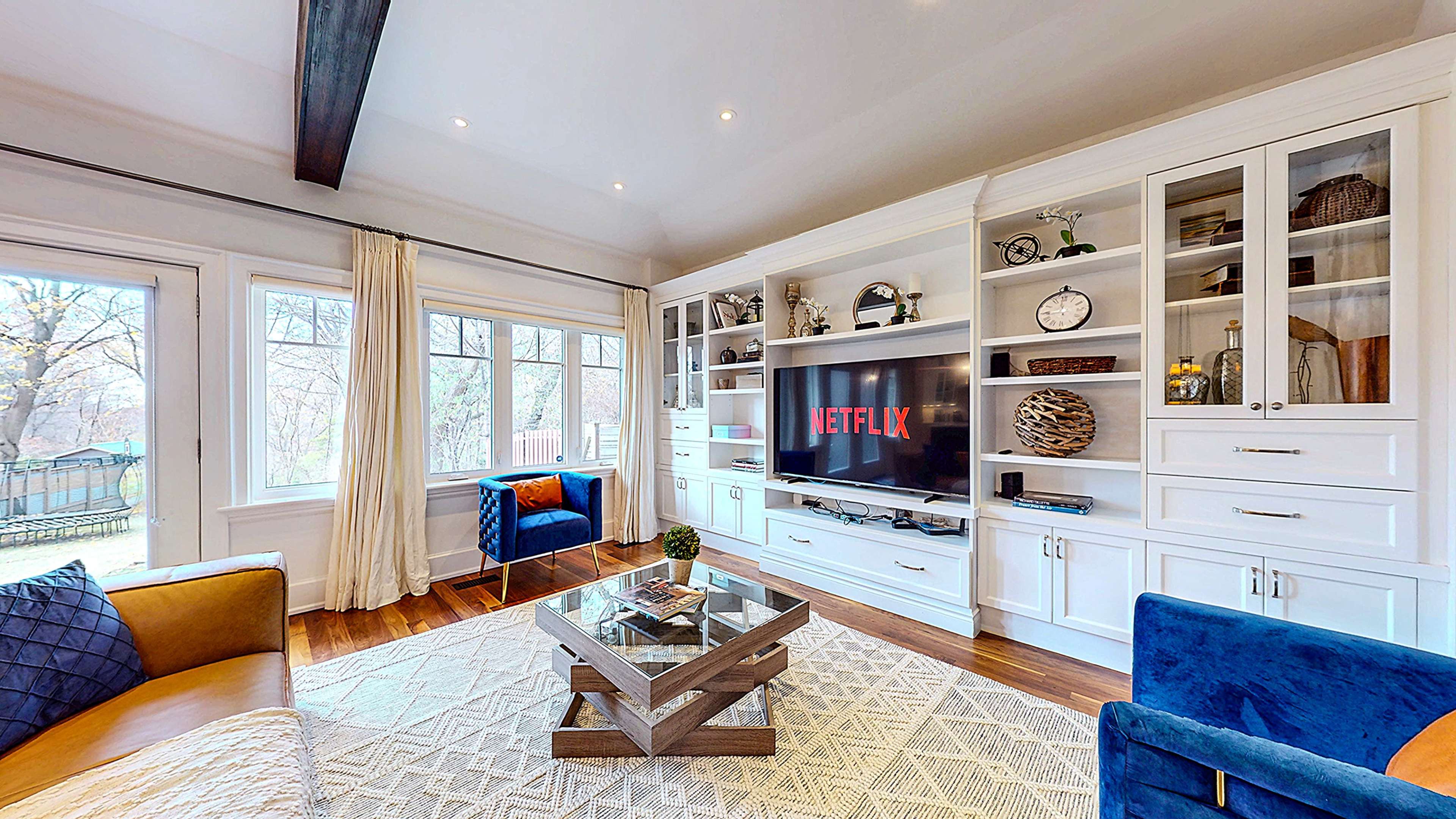 The living room features a television displaying the Netflix logo, surrounded by built-in shelves, a coffee table, and seating areas with blue and beige sofas.