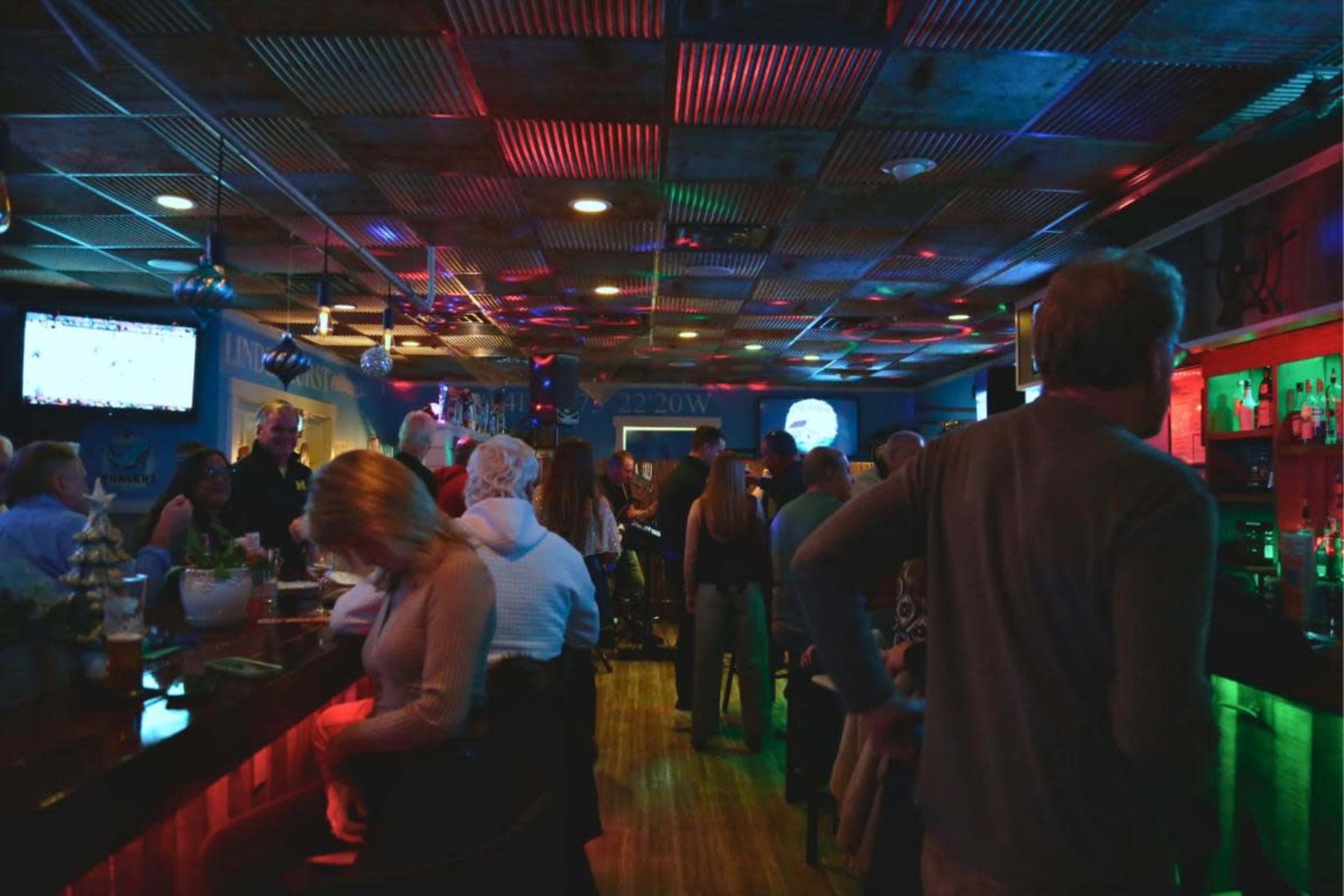 A lively bar scene is filled with people socializing, colorful lights reflecting off the walls, and a television displaying a game in the background.