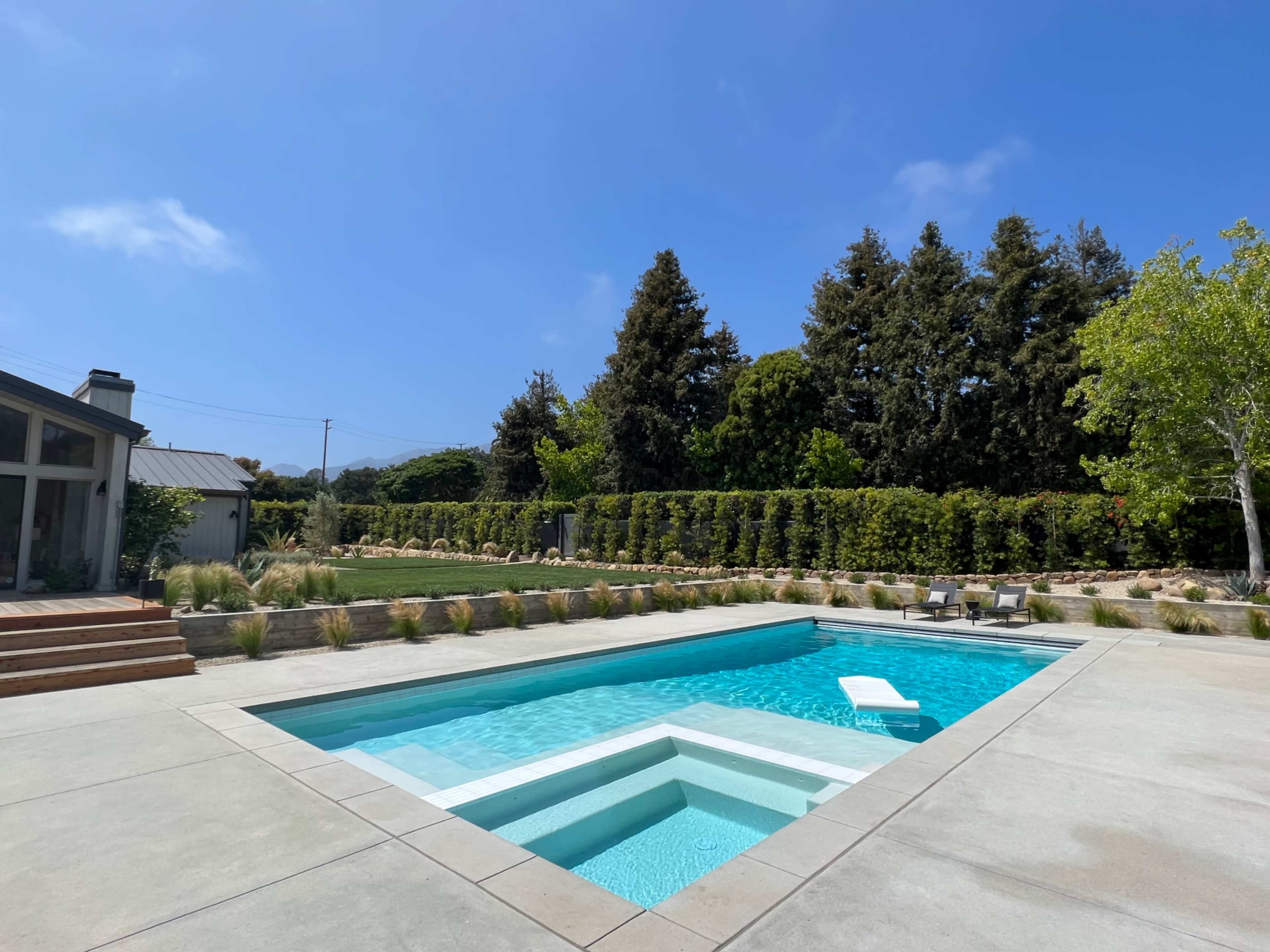 A modern swimming pool with a dive platform is set in a landscaped backyard featuring shrubs and trees under a clear blue sky.