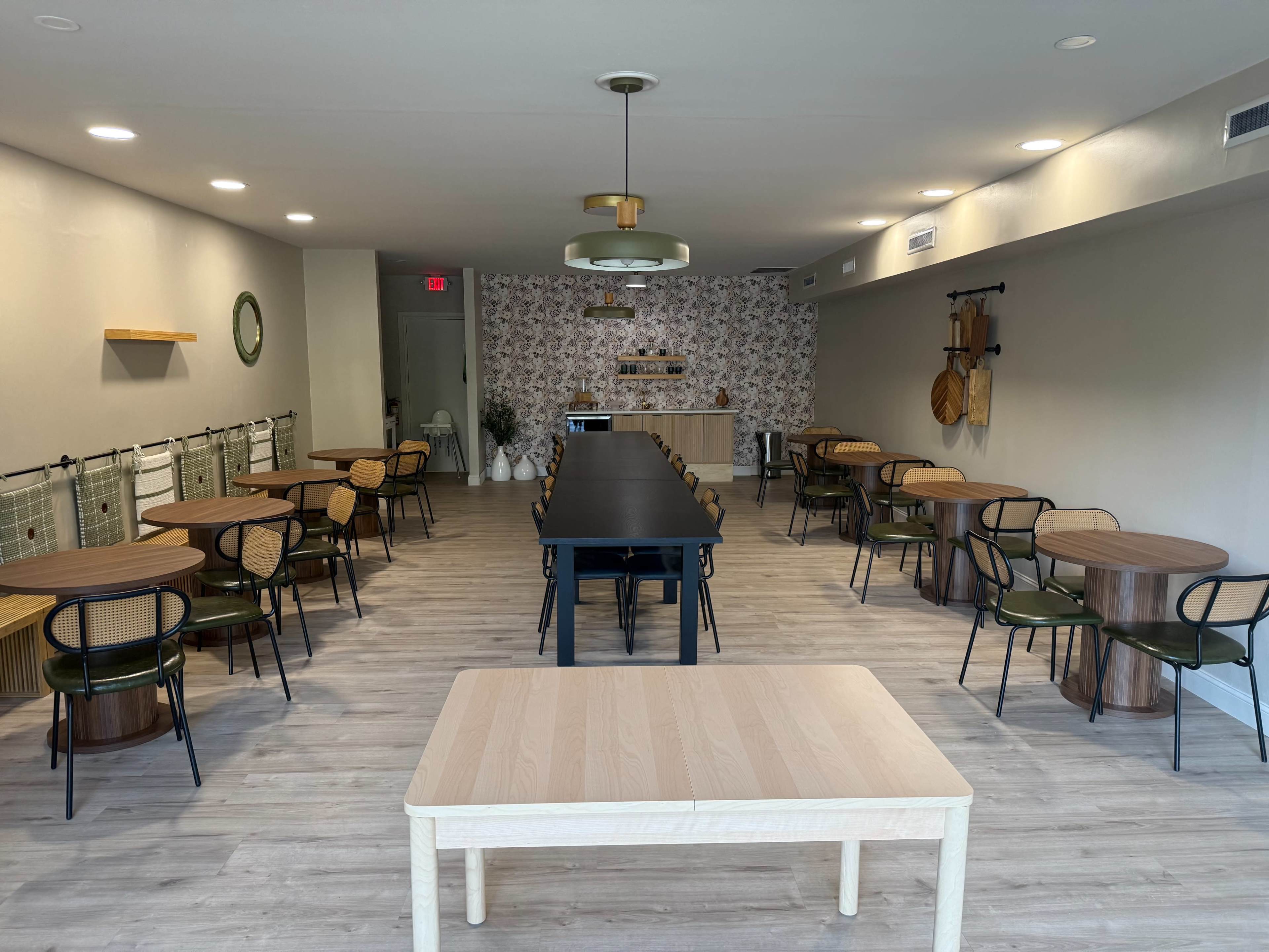 The image shows a spacious dining area with multiple round tables and chairs arranged neatly, featuring a central long table and patterned walls in the background.