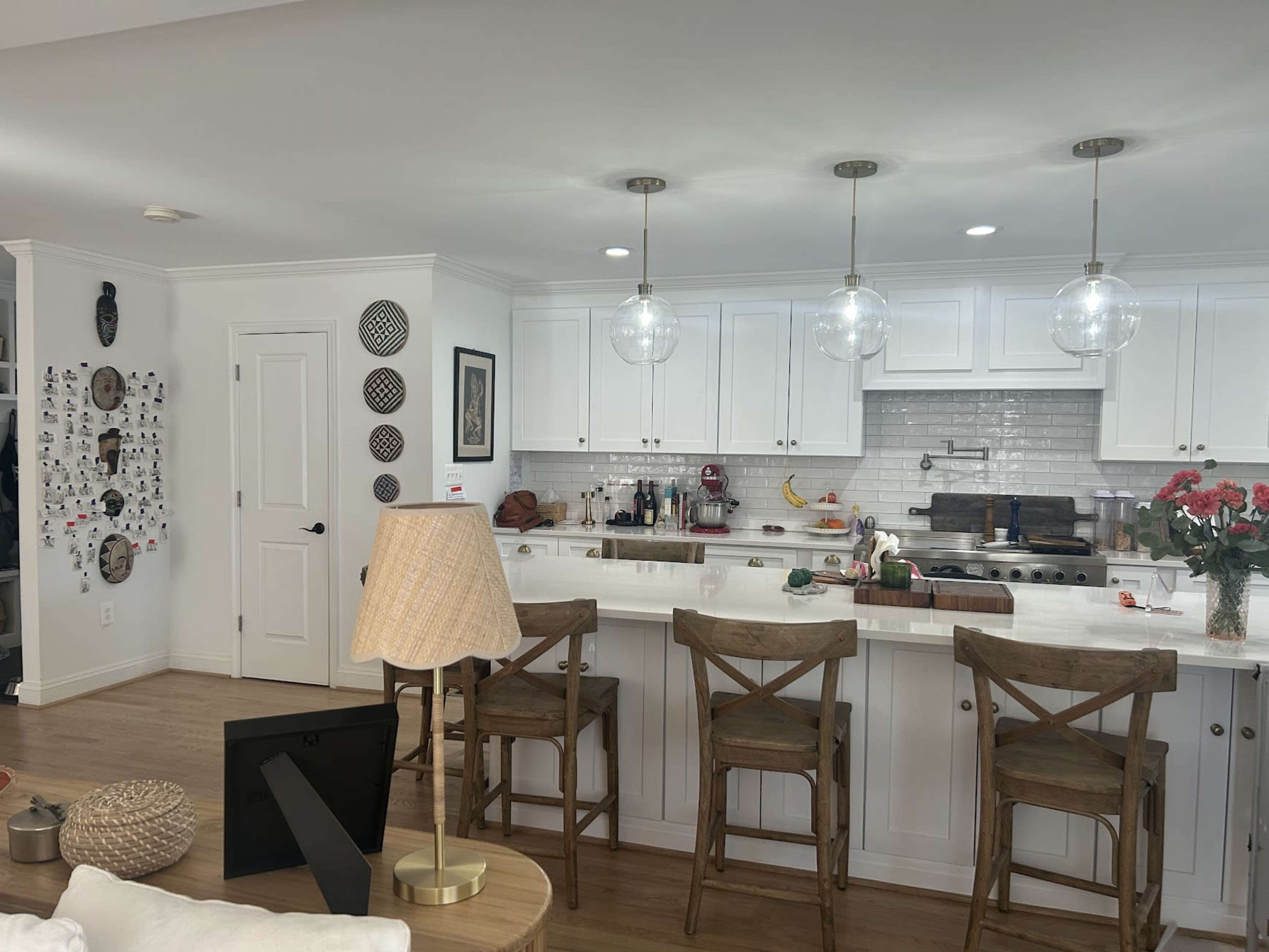 The image shows a modern kitchen with white cabinets, a marble countertop, three pendant lights, and a wooden dining table with four chairs in a brightly lit open space.