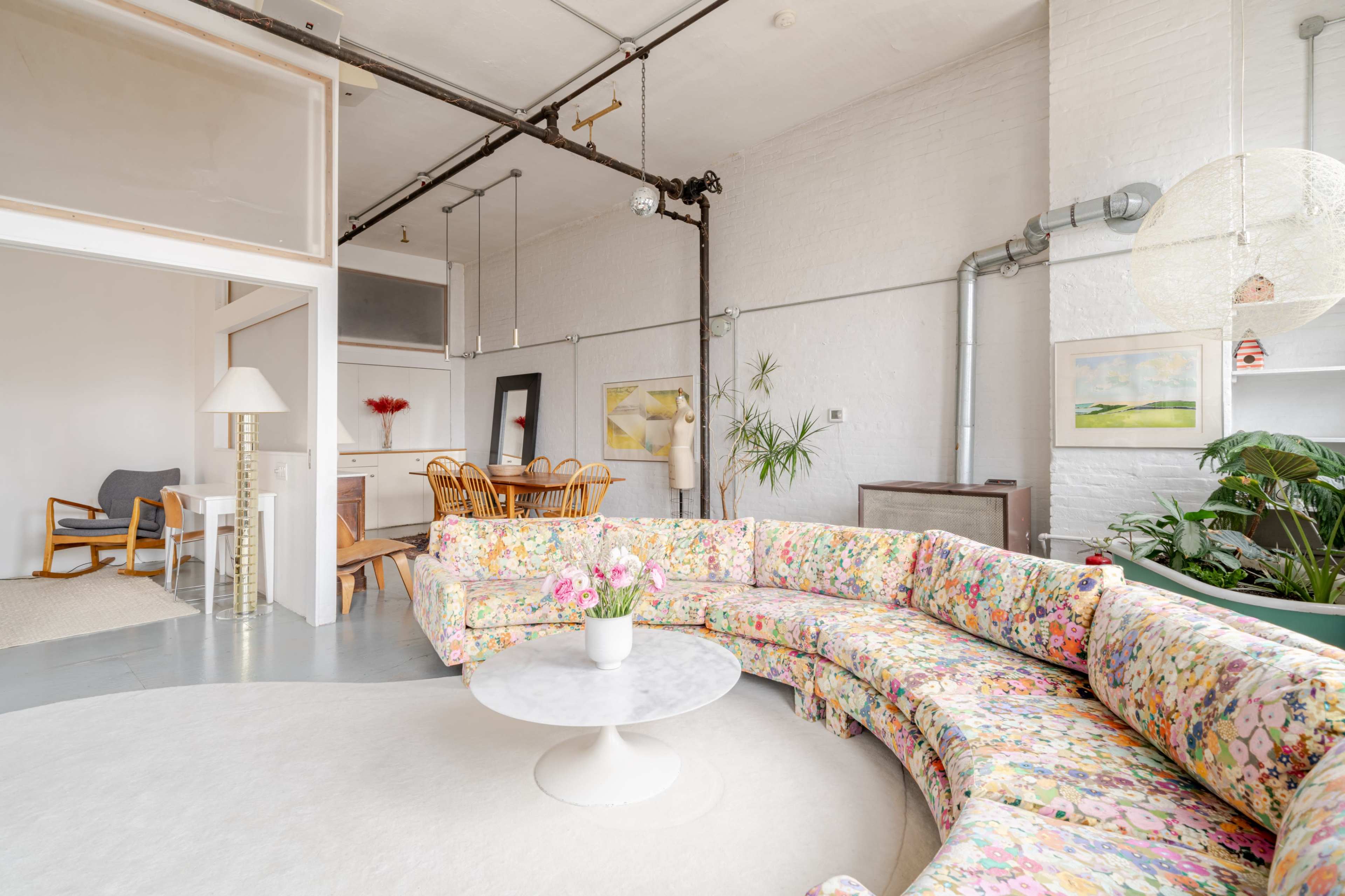 A bright, modern living space features a floral-patterned curved sofa, a round marble coffee table, and dining area with wooden chairs, set against white brick walls and exposed industrial details.