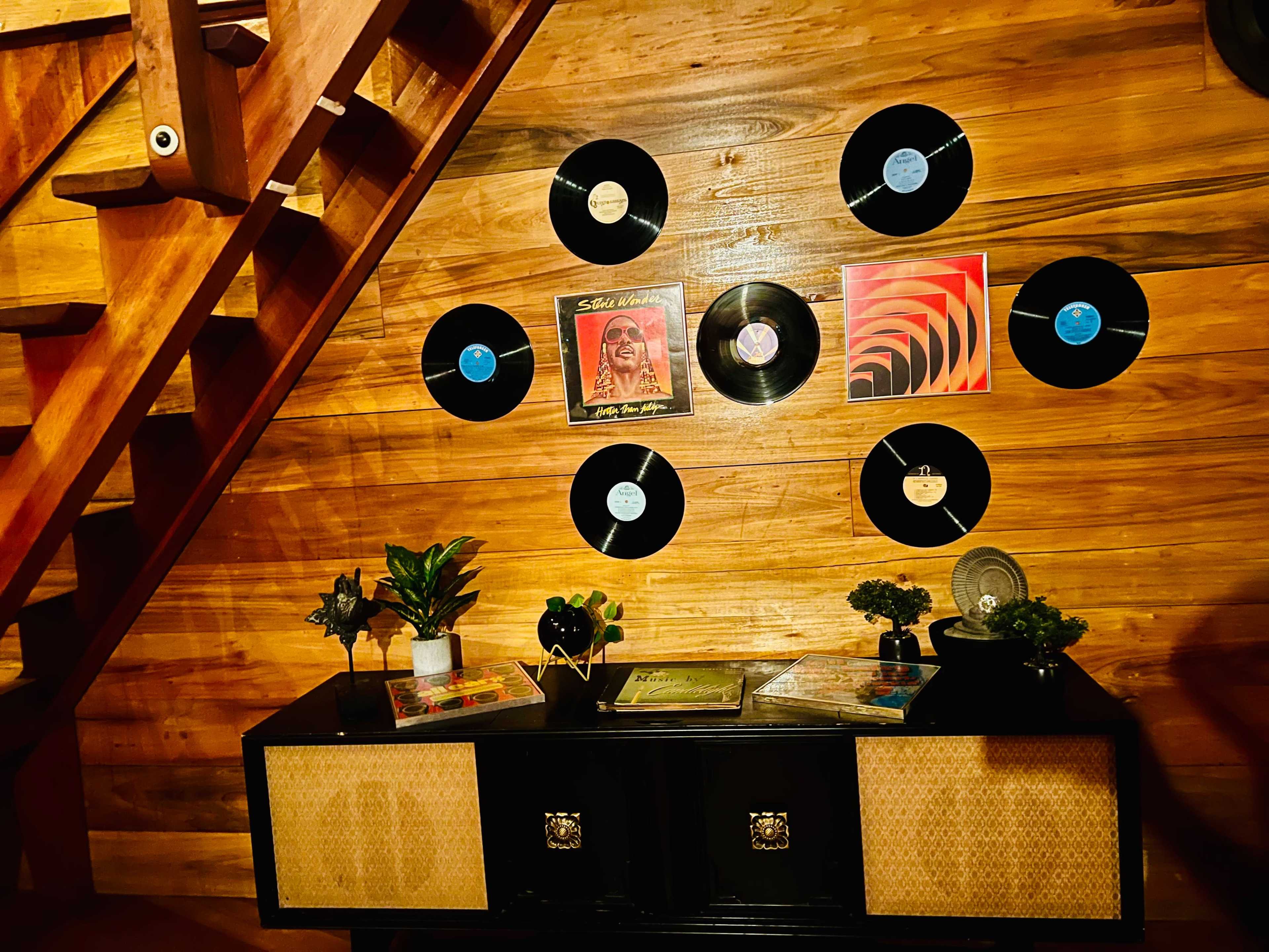 A wooden staircase leads to a wall adorned with records above a black cabinet, which is decorated with small plants and various magazines.