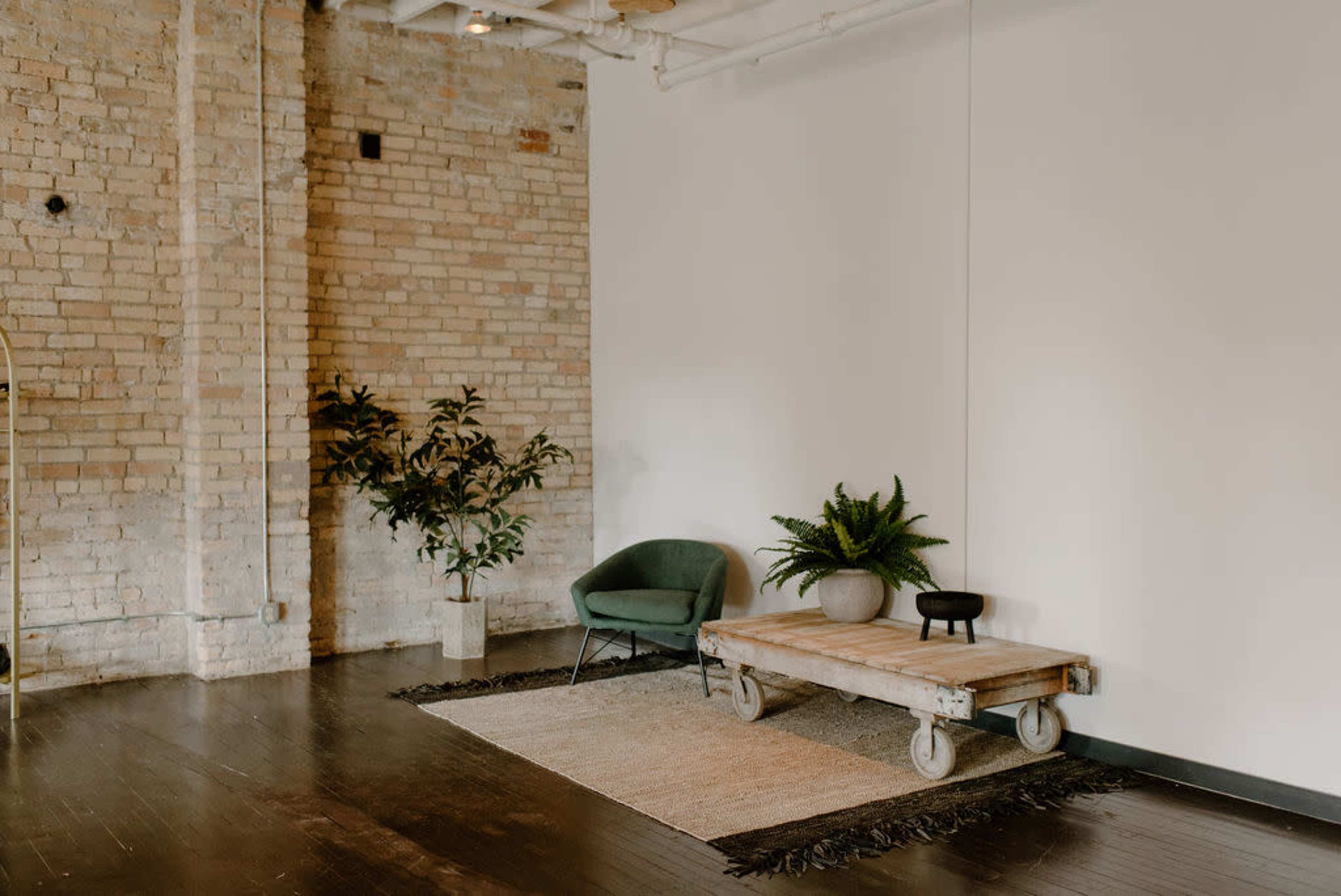 A minimalistic room with exposed brick walls, a green chair, a wooden table on wheels, and decorative plants on the floor.