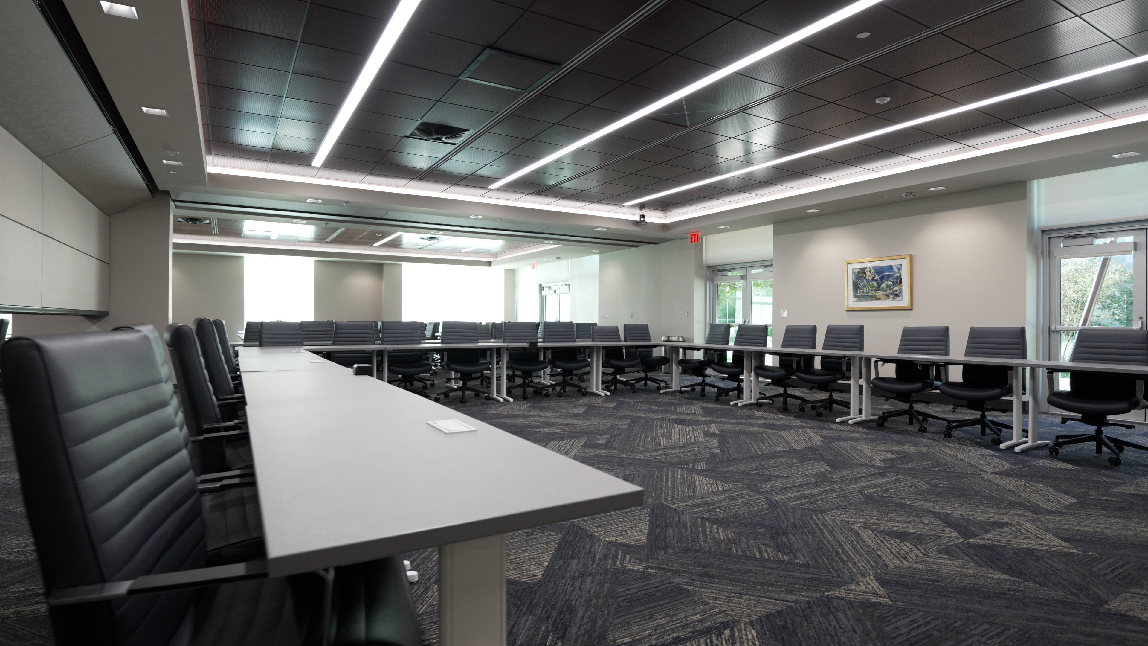 A modern conference room with a long rectangular table, multiple office chairs, and large windows providing natural light.