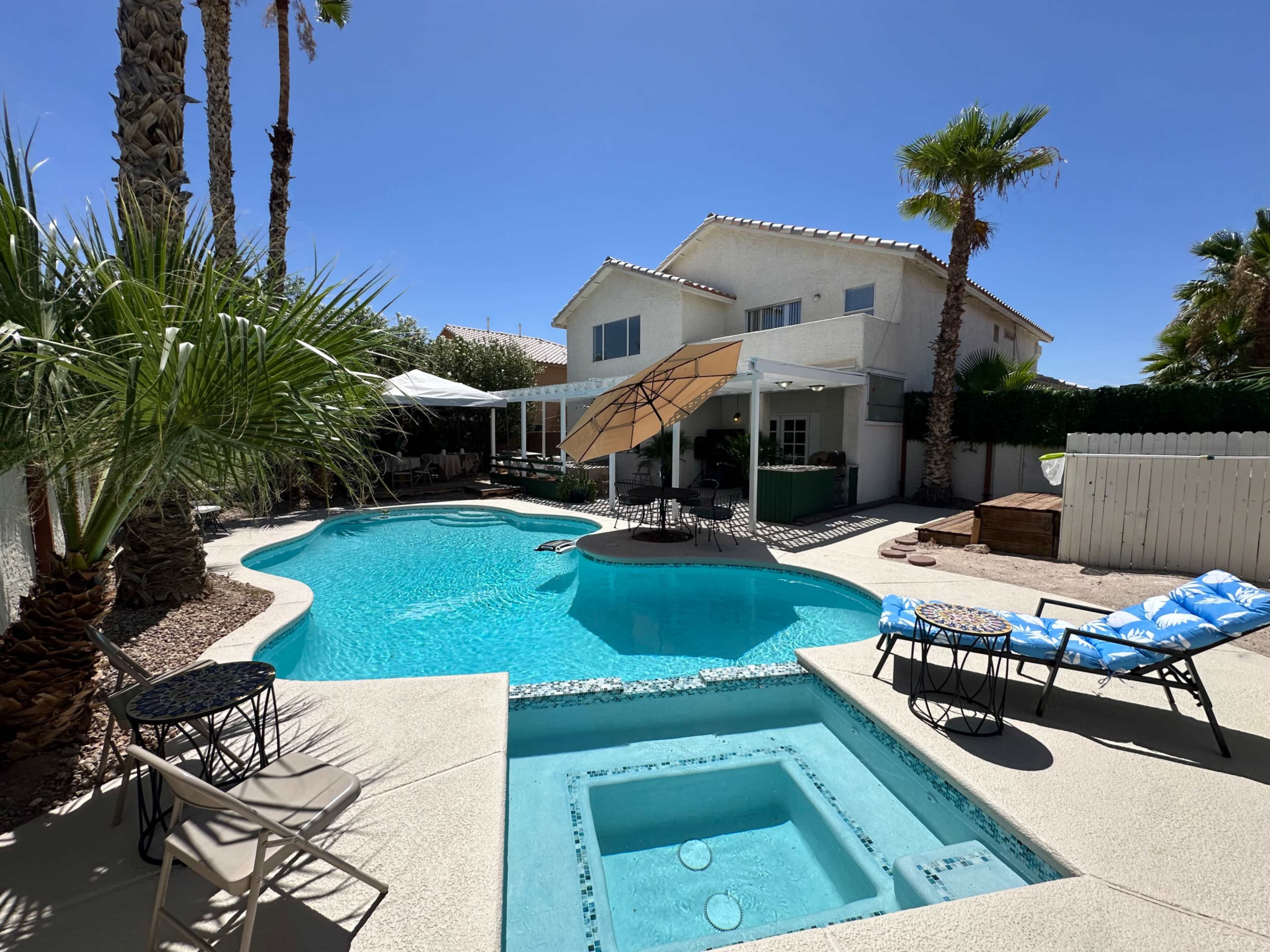 The image shows a backyard with a swimming pool, a spa, several lounge chairs, and a house with a patio surrounded by palm trees.