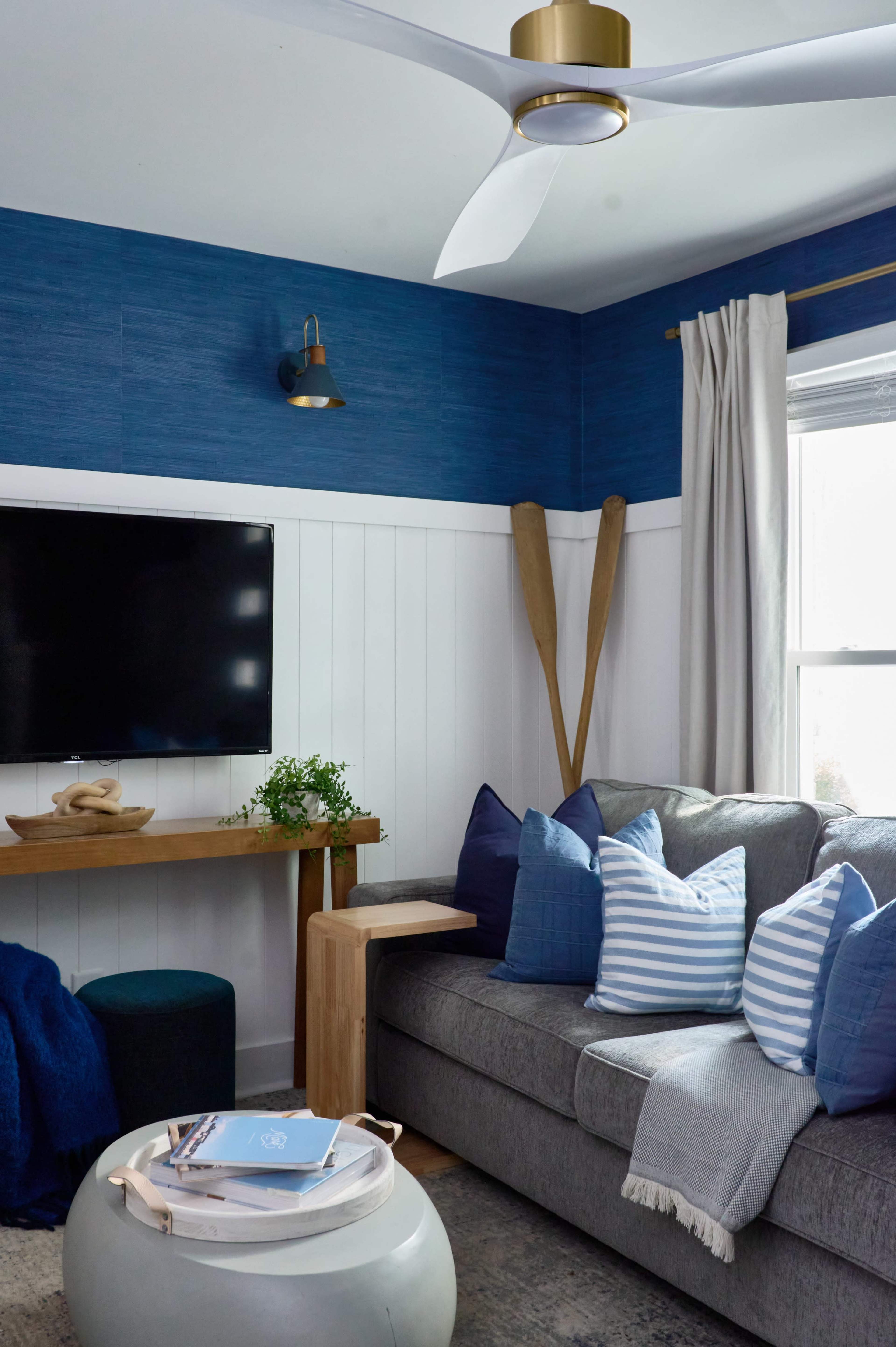 The image shows a cozy living room with a gray couch adorned with blue and white striped pillows, a wooden table, a modern ceiling fan, and a wall-mounted television.