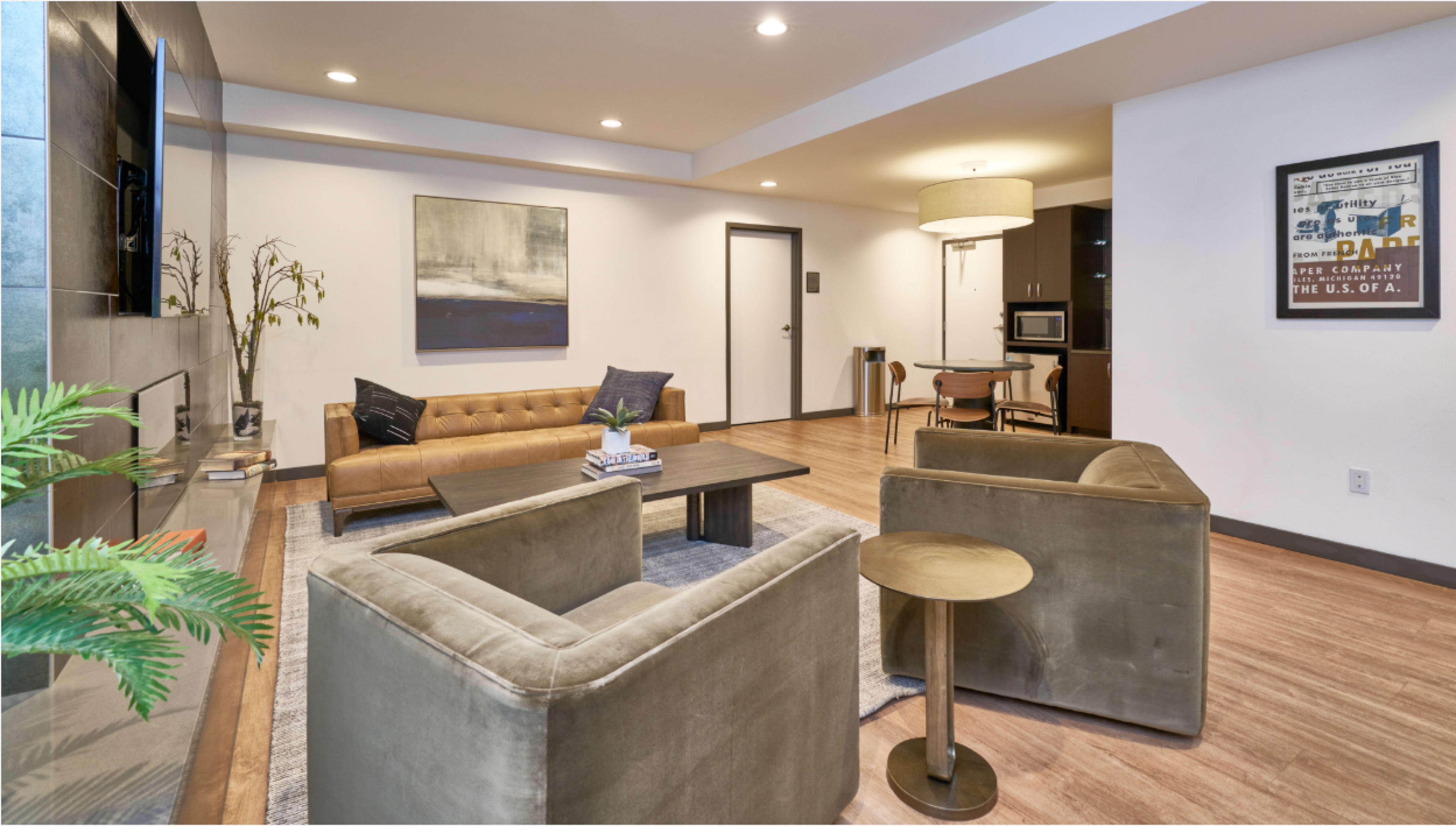 The image shows a modern living room with a brown leather sofa, two gray chairs, a coffee table, and an adjacent kitchenette.