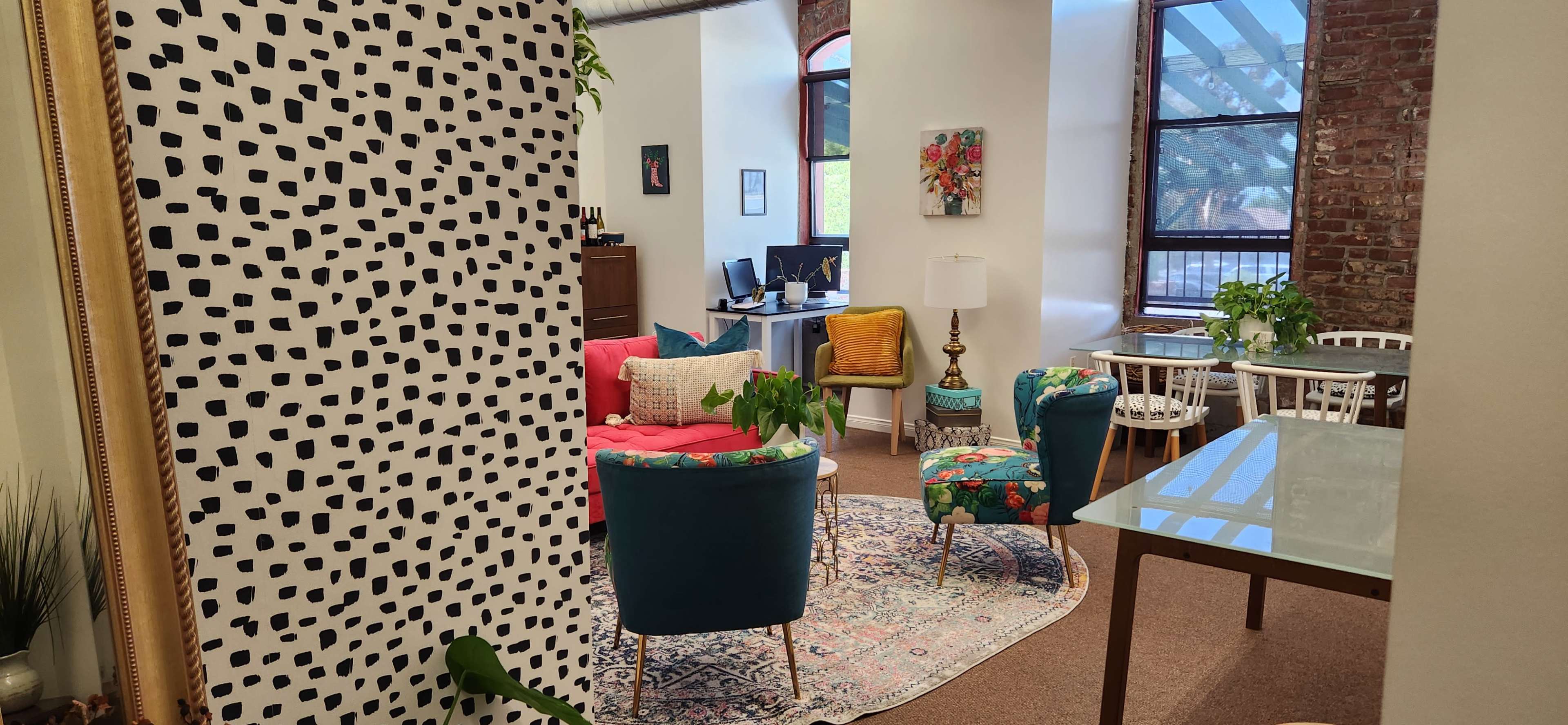 A modern office space featuring colorful seating, a large area rug, and workstations in a well-lit room with brick walls and large windows.