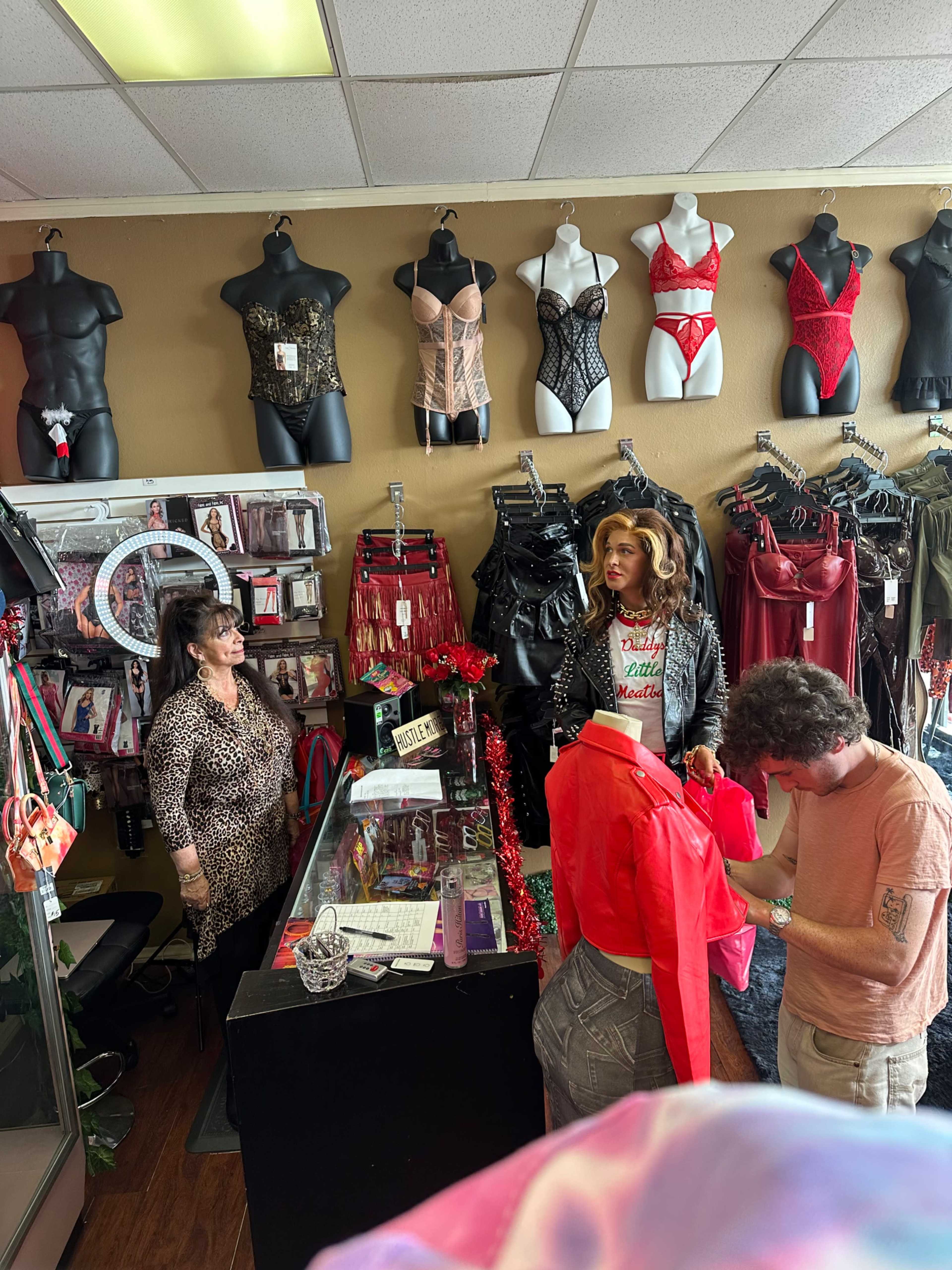 A woman stands behind a counter in a lingerie store while a man examines a red garment and a mannequin in lingerie is displayed nearby.