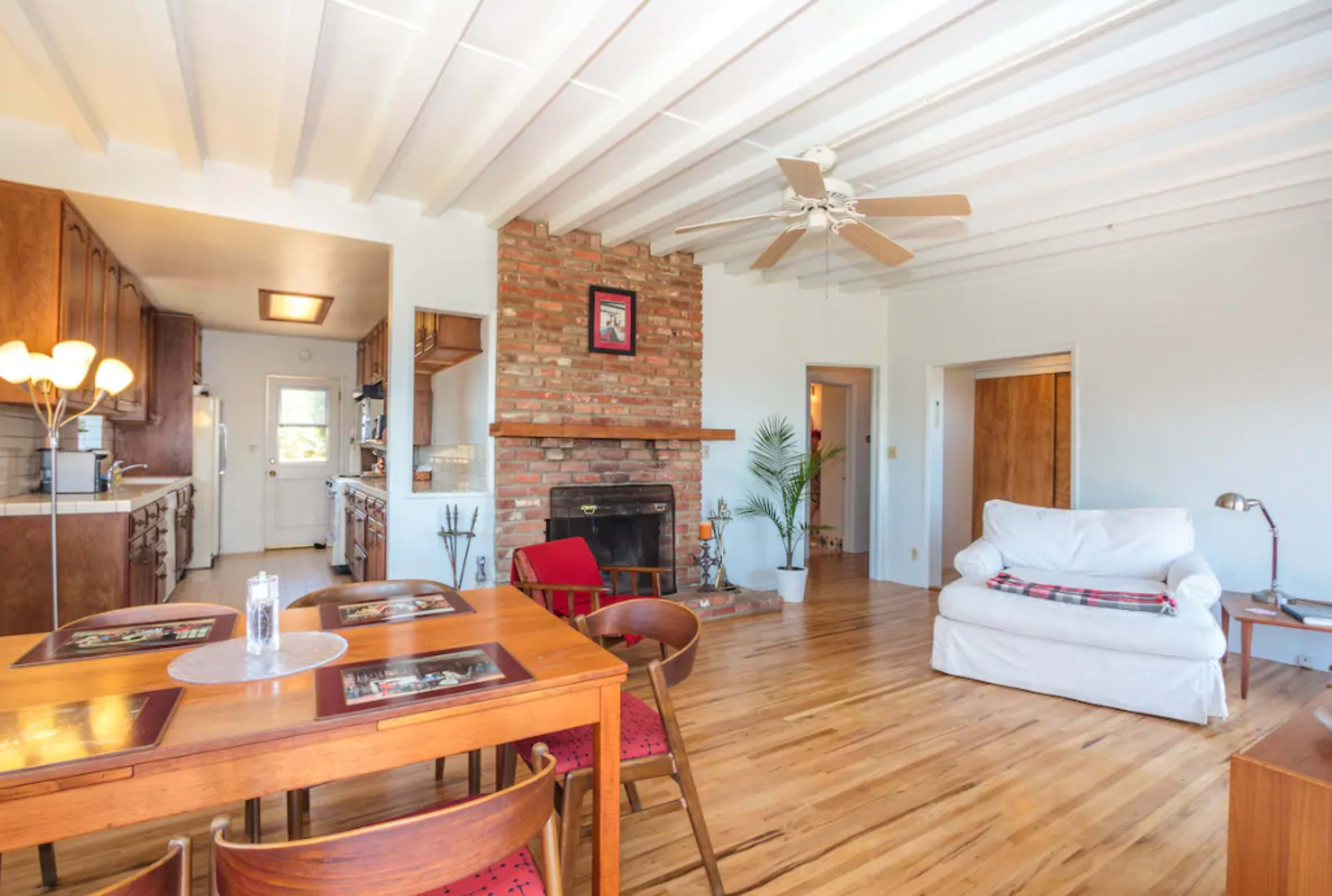 A spacious living area with a dining table, a couch, a brick fireplace, and a ceiling fan, complemented by wooden floors and simple decor.