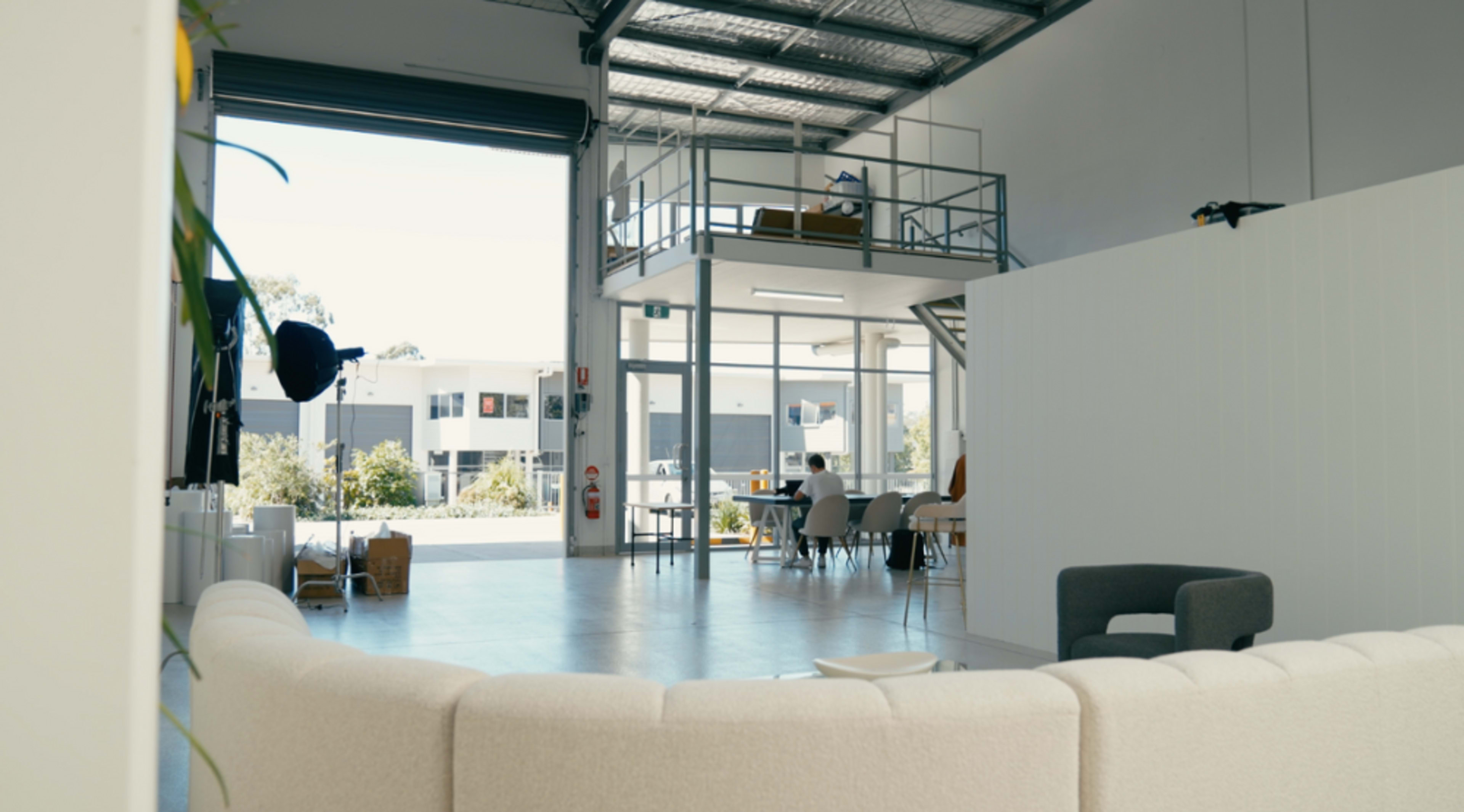 An open, modern workspace featuring a seating area, tables, and a second floor visible through large windows.