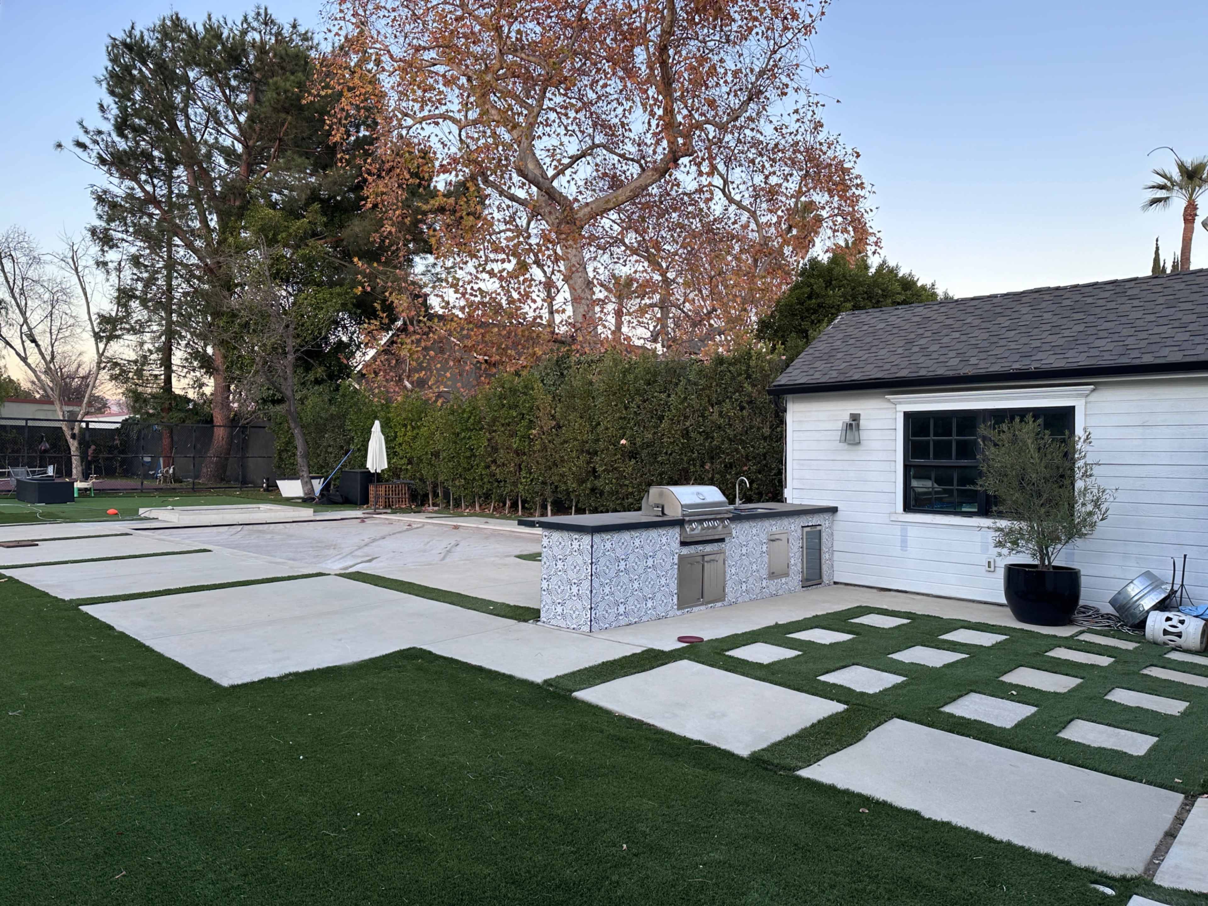 The image shows a backyard with a landscaped area featuring a built-in outdoor grill, stone pathways, and patches of artificial grass.