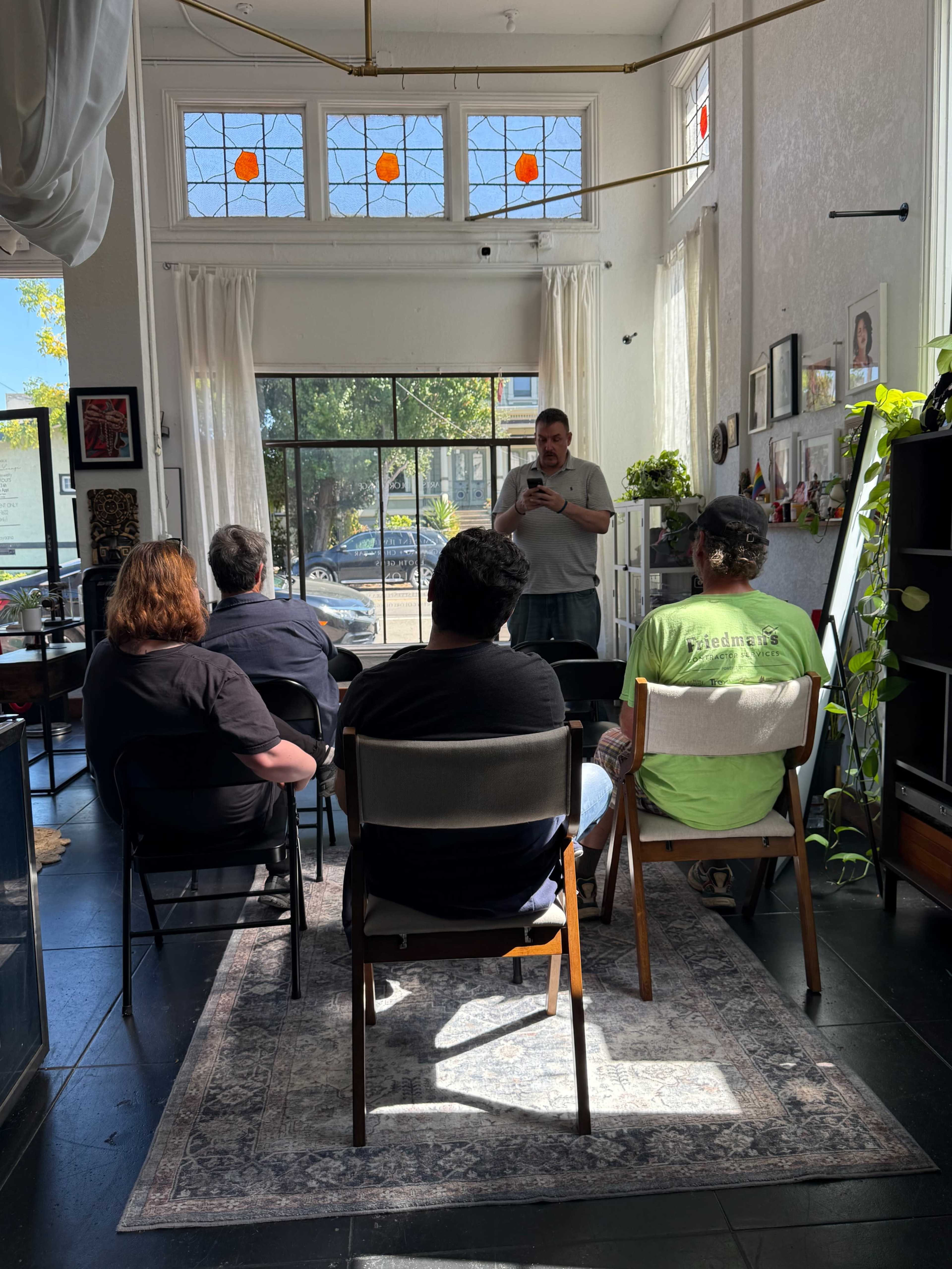A speaker stands in front of an audience seated in a brightly lit room with plants and artworks on the walls.