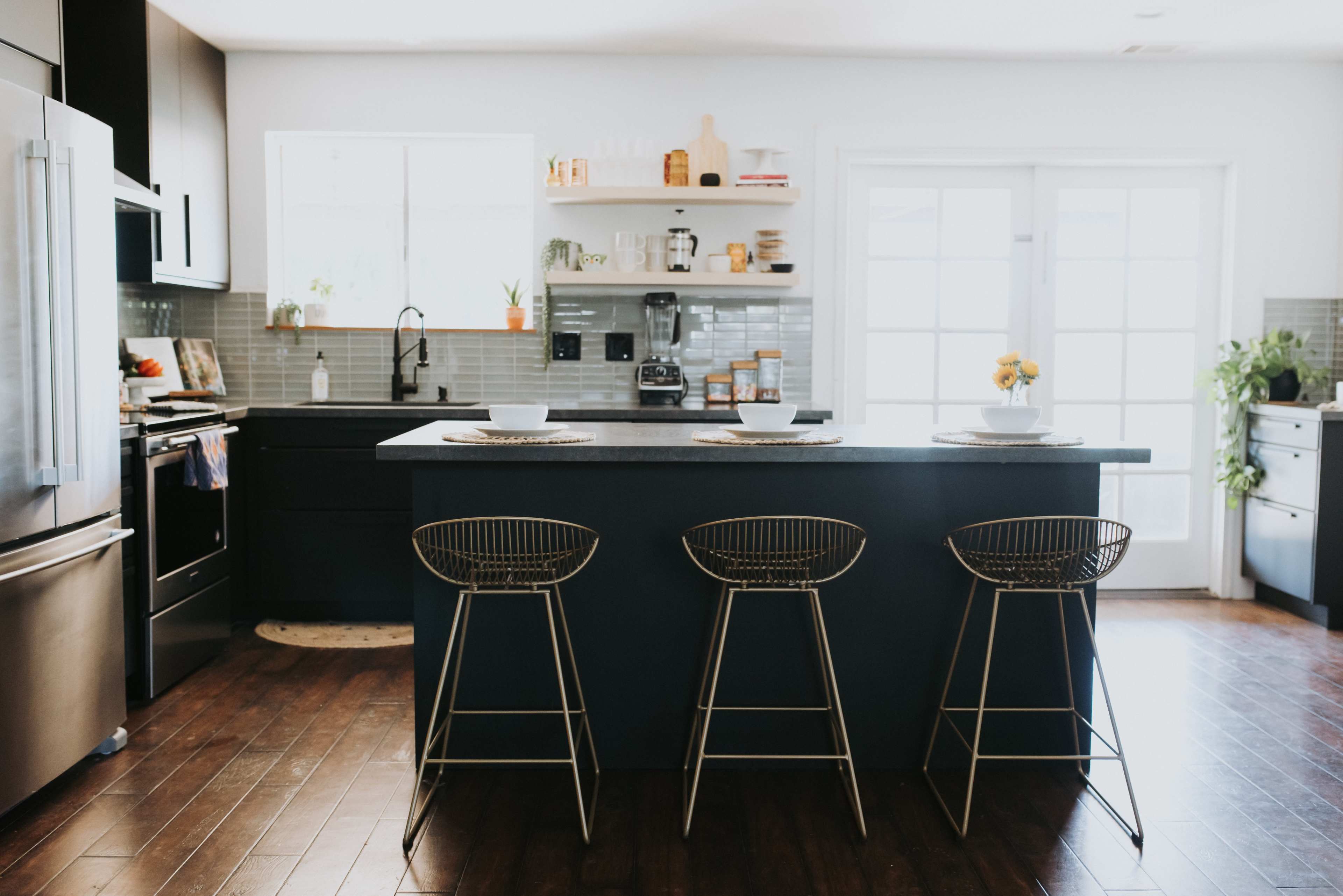 A modern kitchen features a central dark island with three metal bar stools, stainless steel appliances, and open shelving displaying kitchenware.