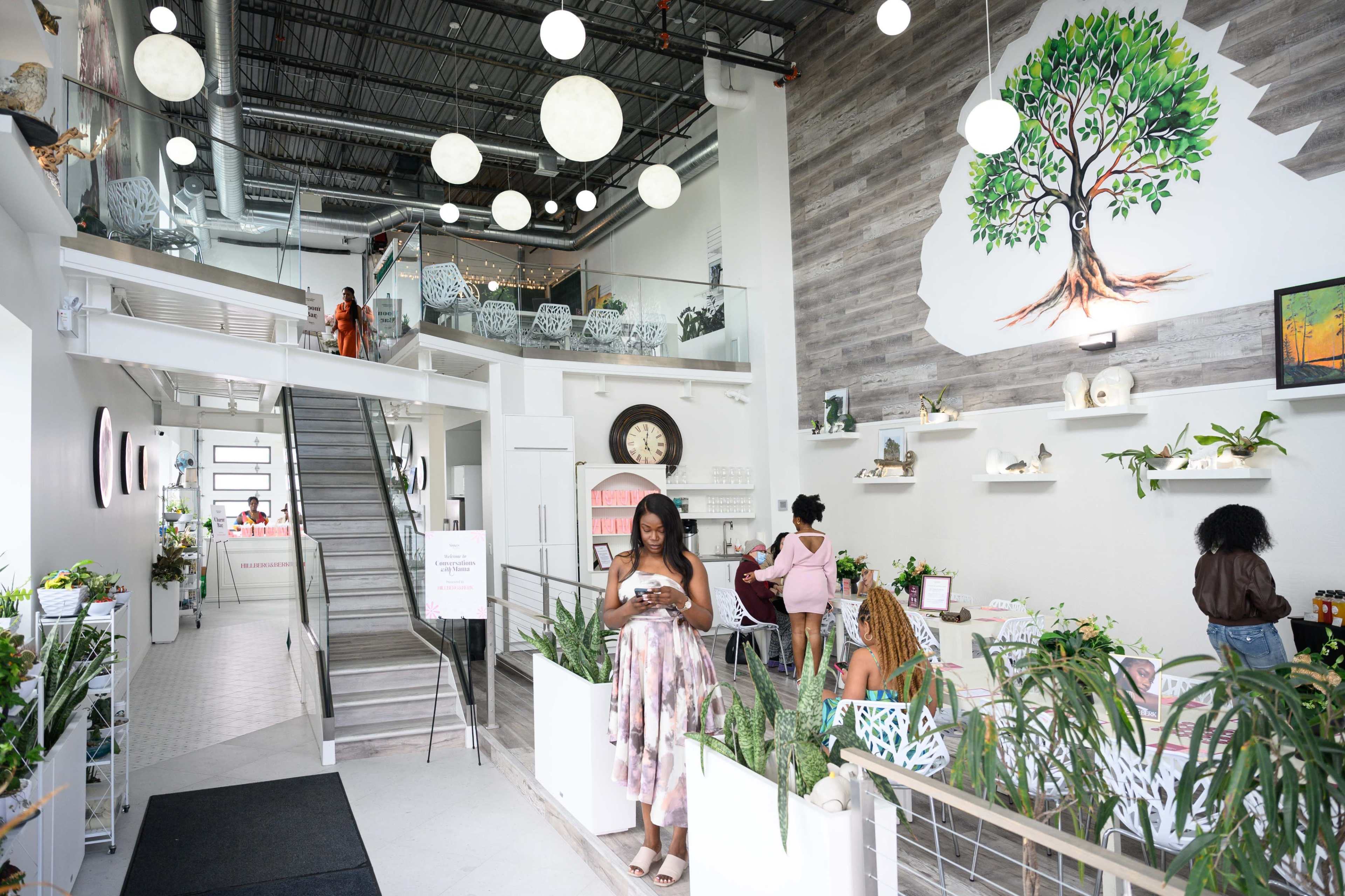 A woman stands in a bright, modern store filled with plants and decorative items, while others browse and interact in the background.
