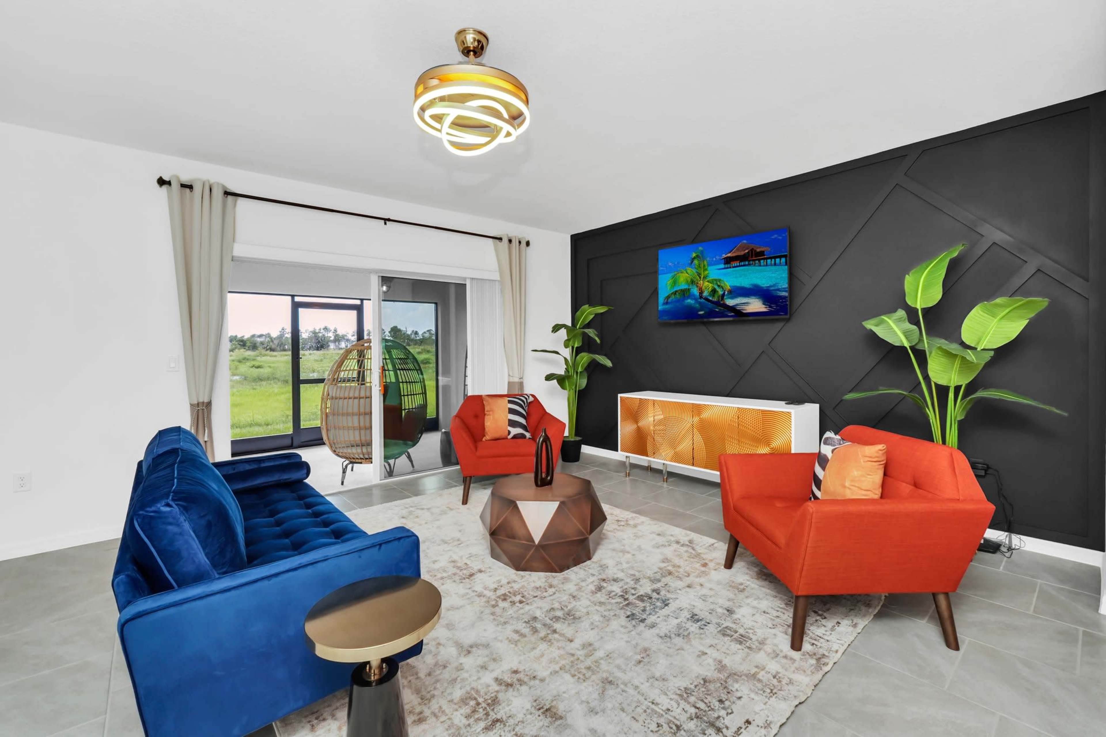 The living room features two orange chairs and a blue sofa centered around a geometric coffee table, with a large TV mounted on a dark accent wall and a view of greenery through sliding glass doors.