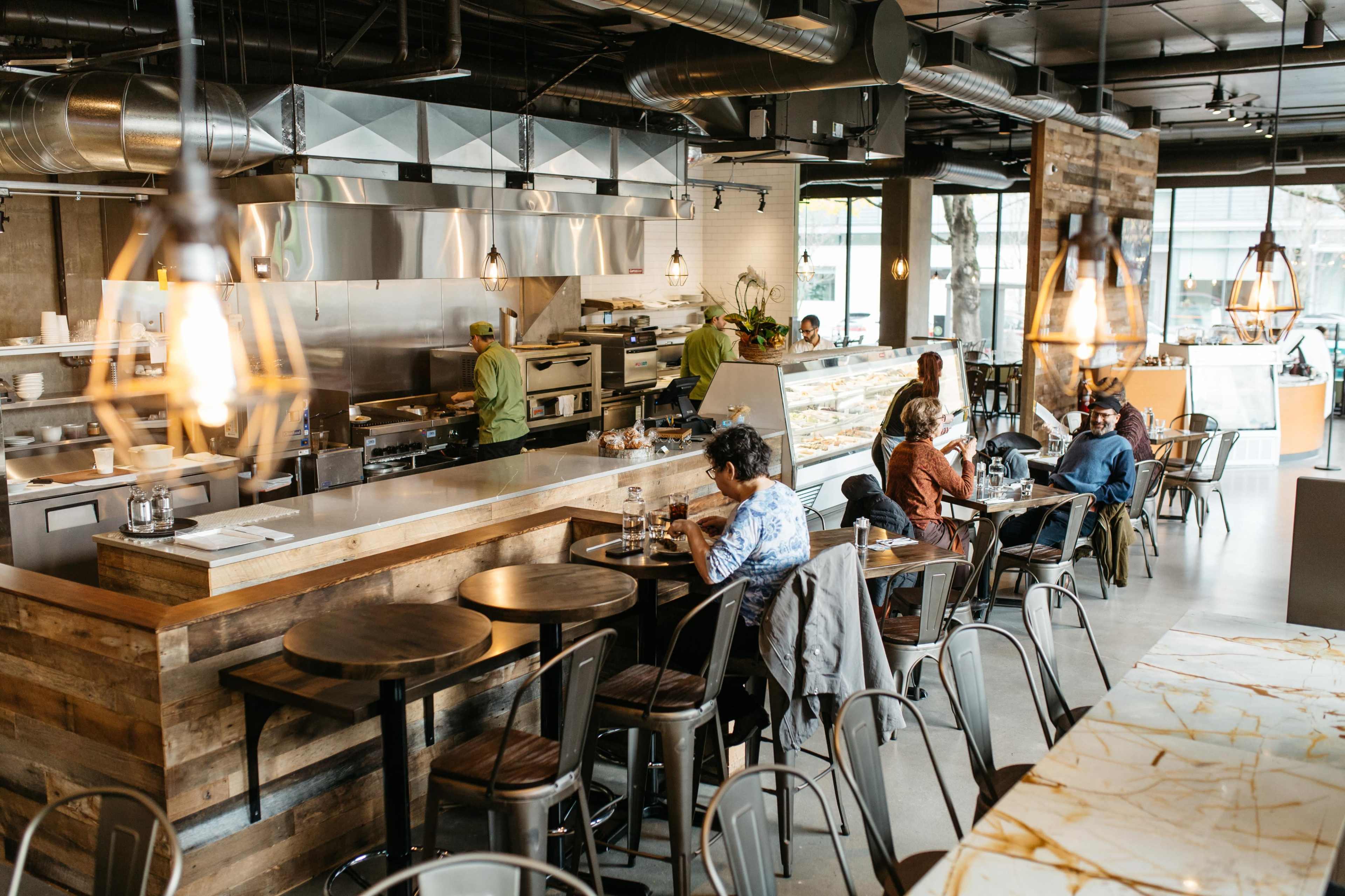 The image shows a modern restaurant interior with diners seated at tables and a visible open kitchen area.