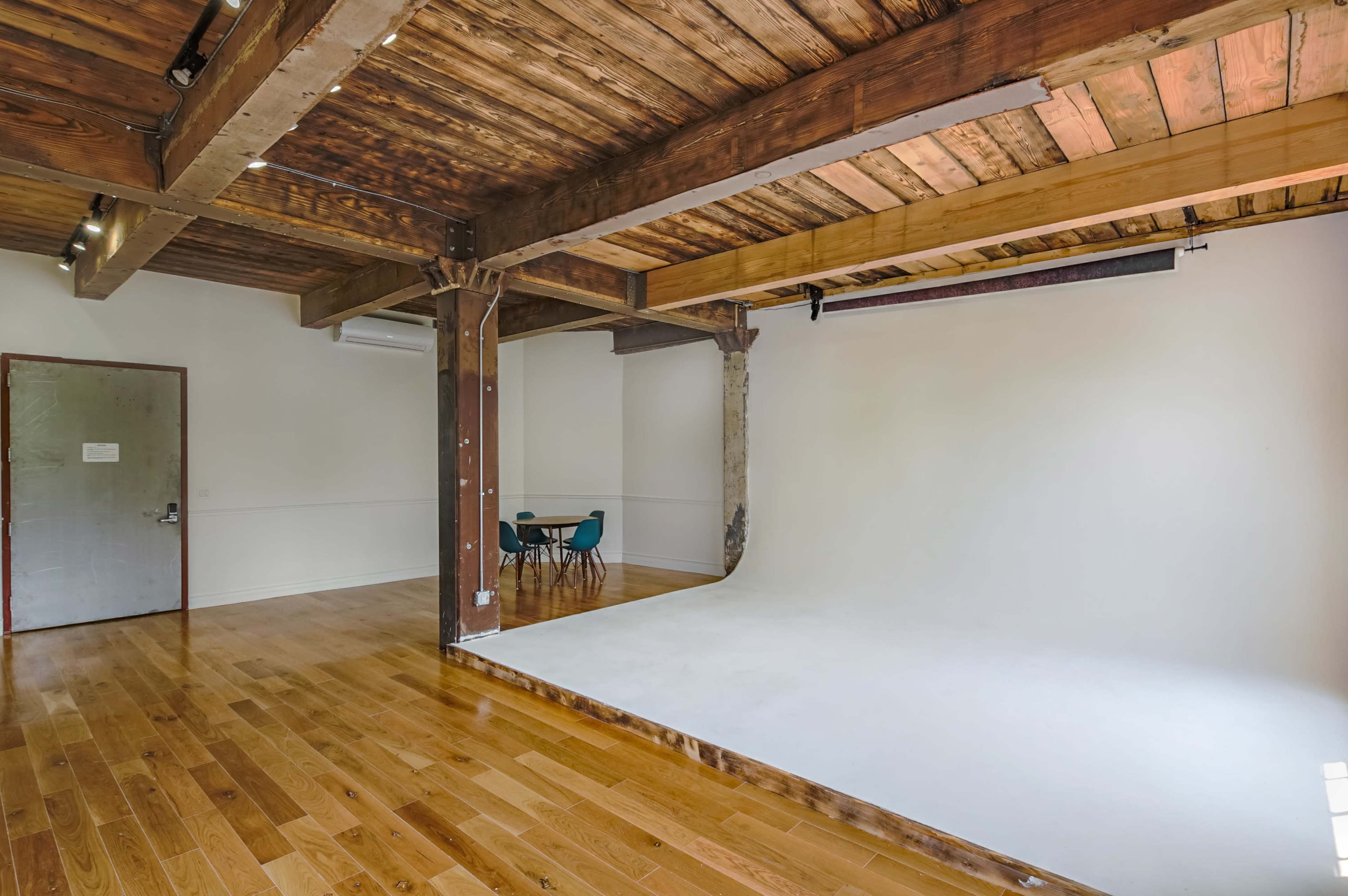 The image shows a spacious interior with wooden beams, a light wooden floor, and a blank backdrop area, along with a small table and chairs in the background.
