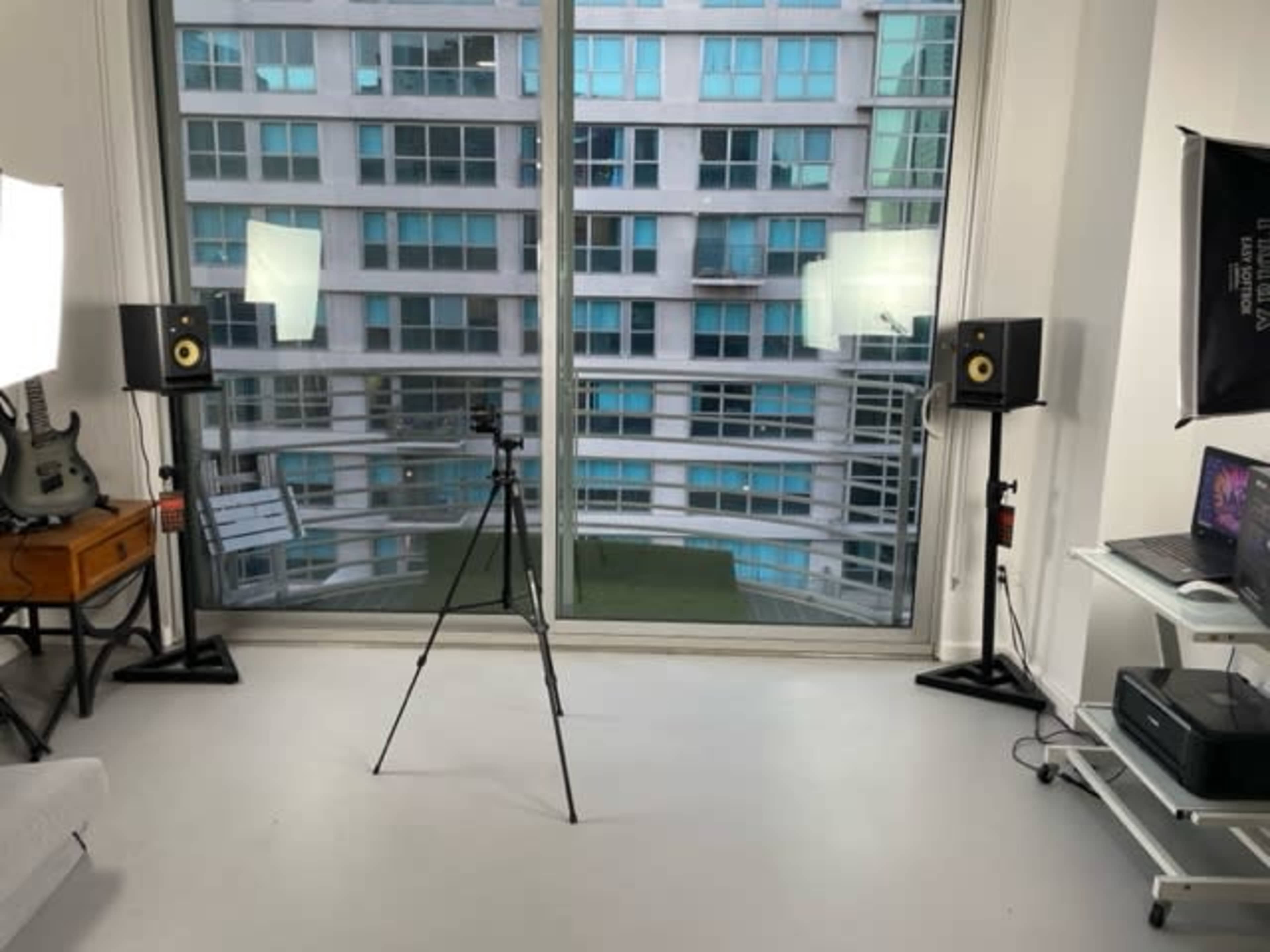 A well-lit room features a tripod in the center, with studio lights positioned nearby and windows showing an exterior view of an apartment building.