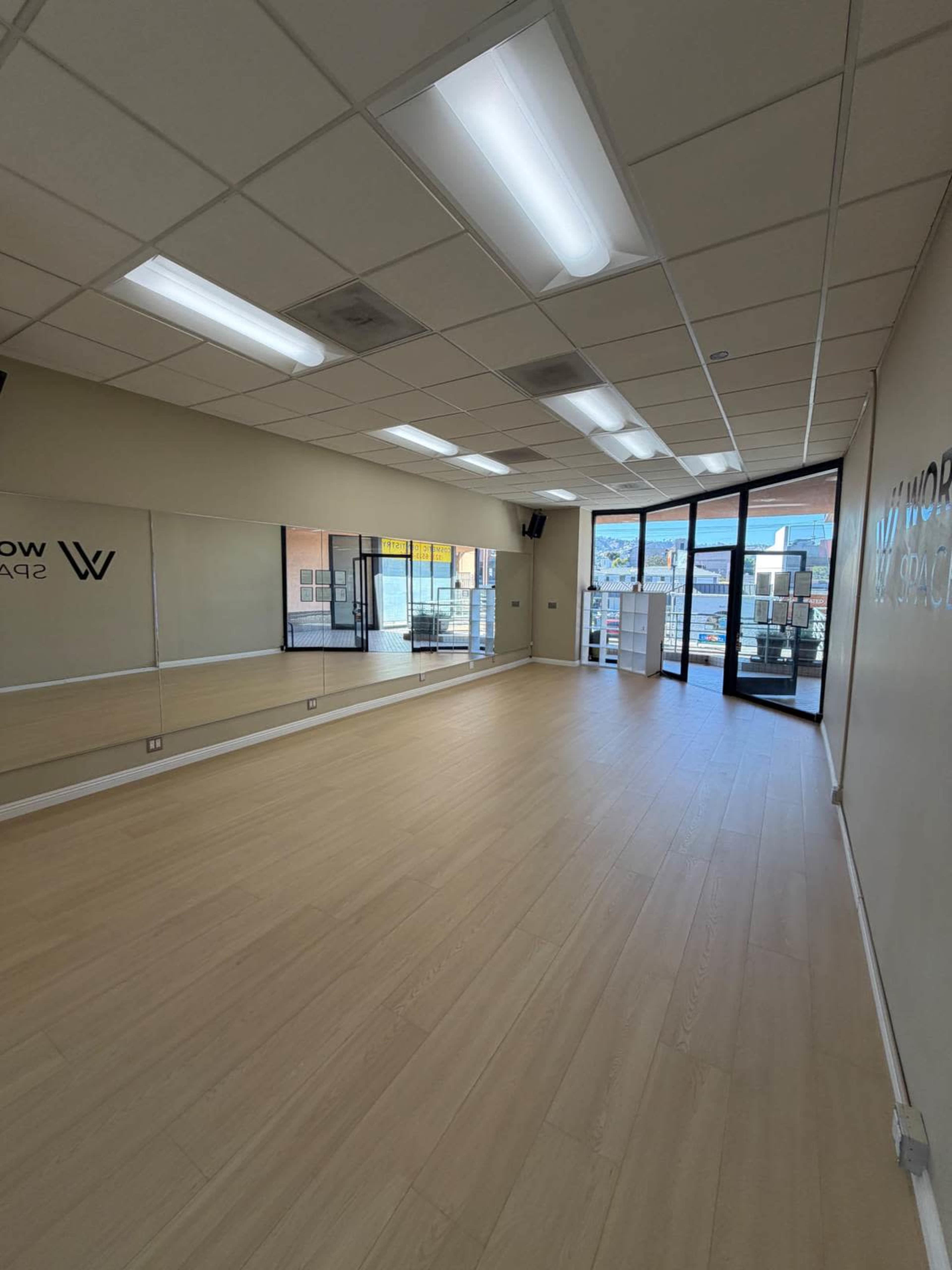 The image shows a spacious studio with wooden flooring, large mirrors on one wall, and windows that provide natural light.