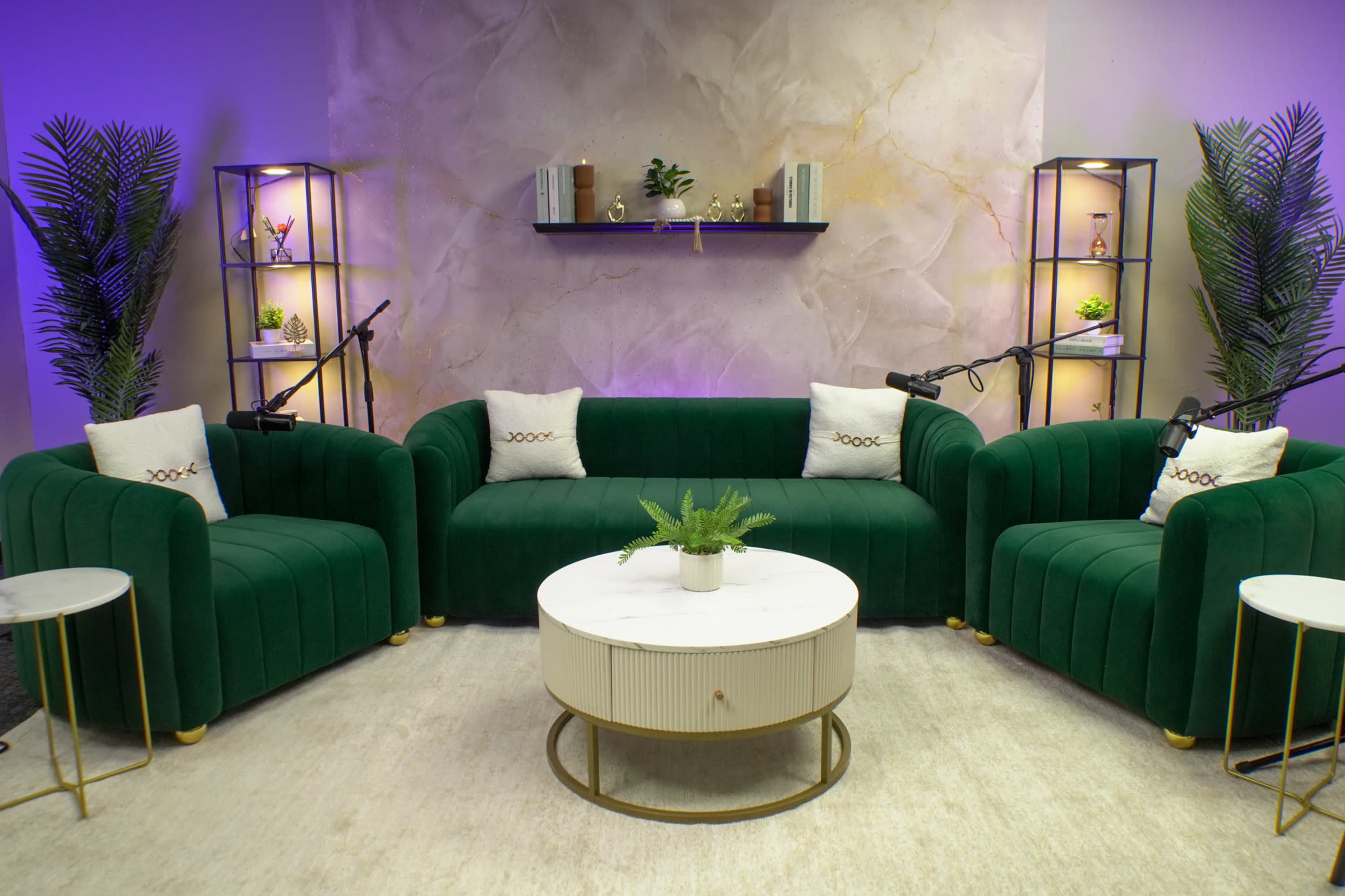 The image shows a modern seating area with two green upholstered sofas, a round coffee table, and decorative shelving units in the background, all arranged in a well-lit space.