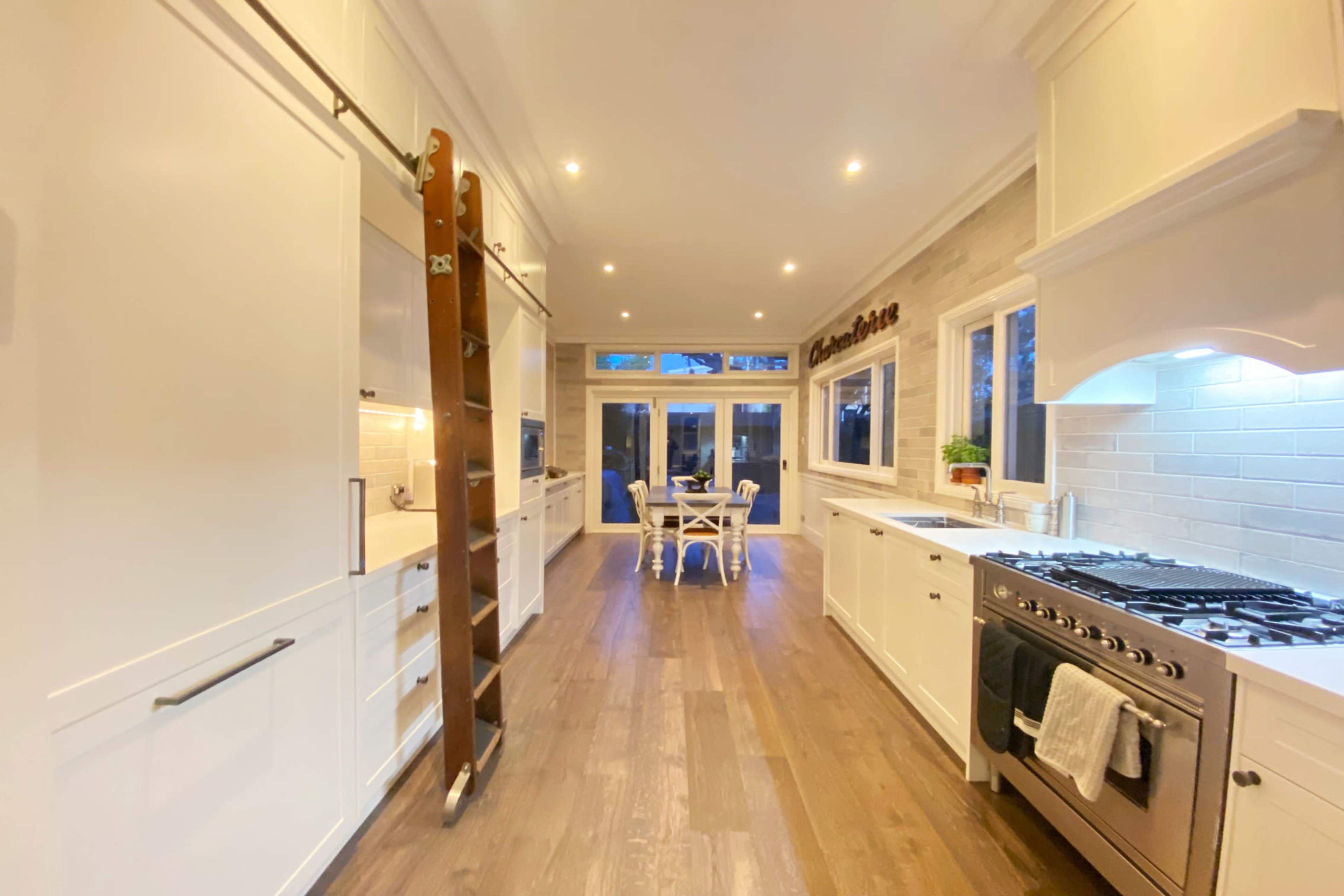 A modern kitchen features white cabinetry, wooden flooring, and a dining area with a table and chairs visible through large windows.
