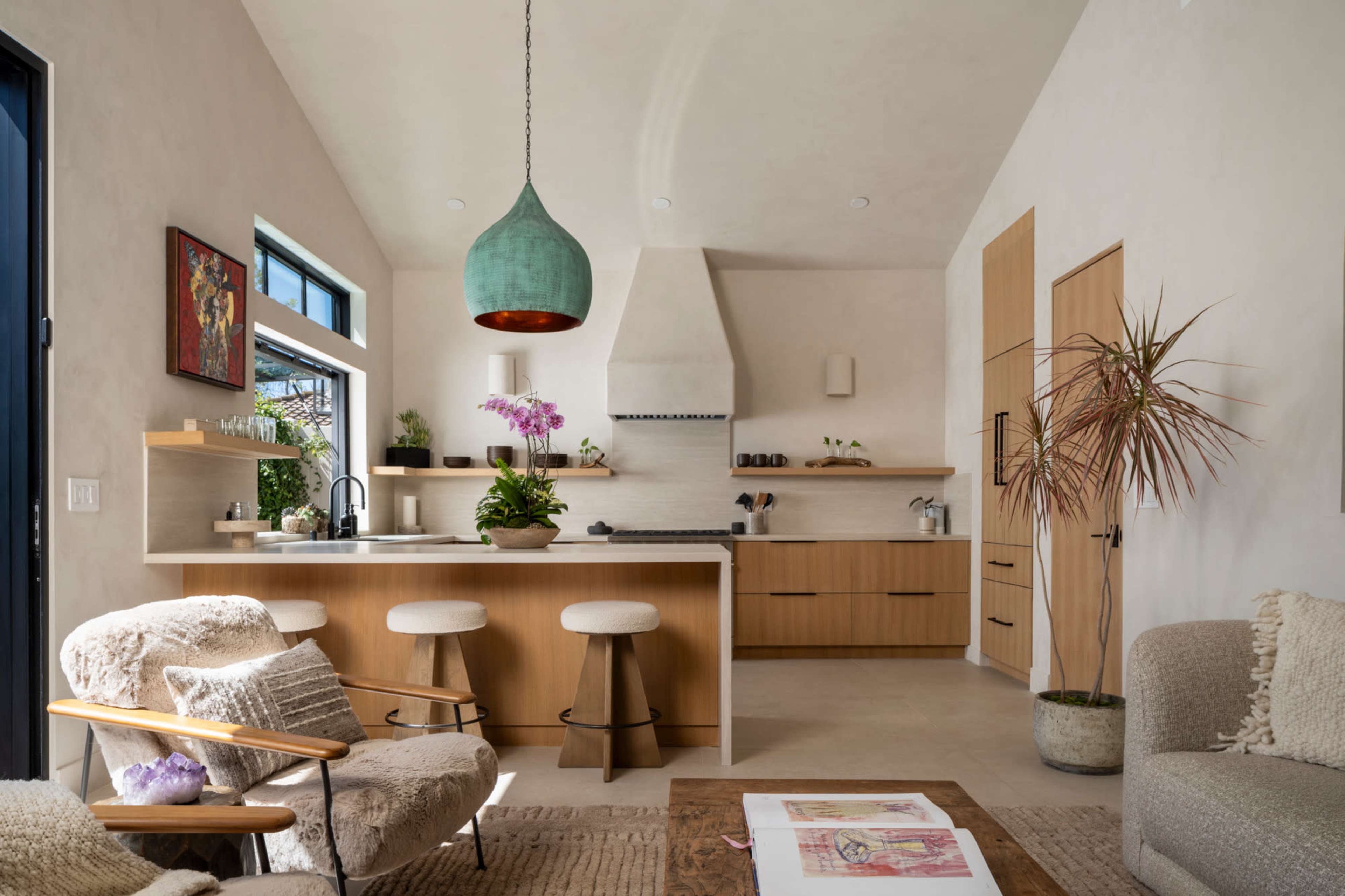 The image shows a modern kitchen and living area featuring wooden cabinetry, a large pendant light, and seating arrangements.