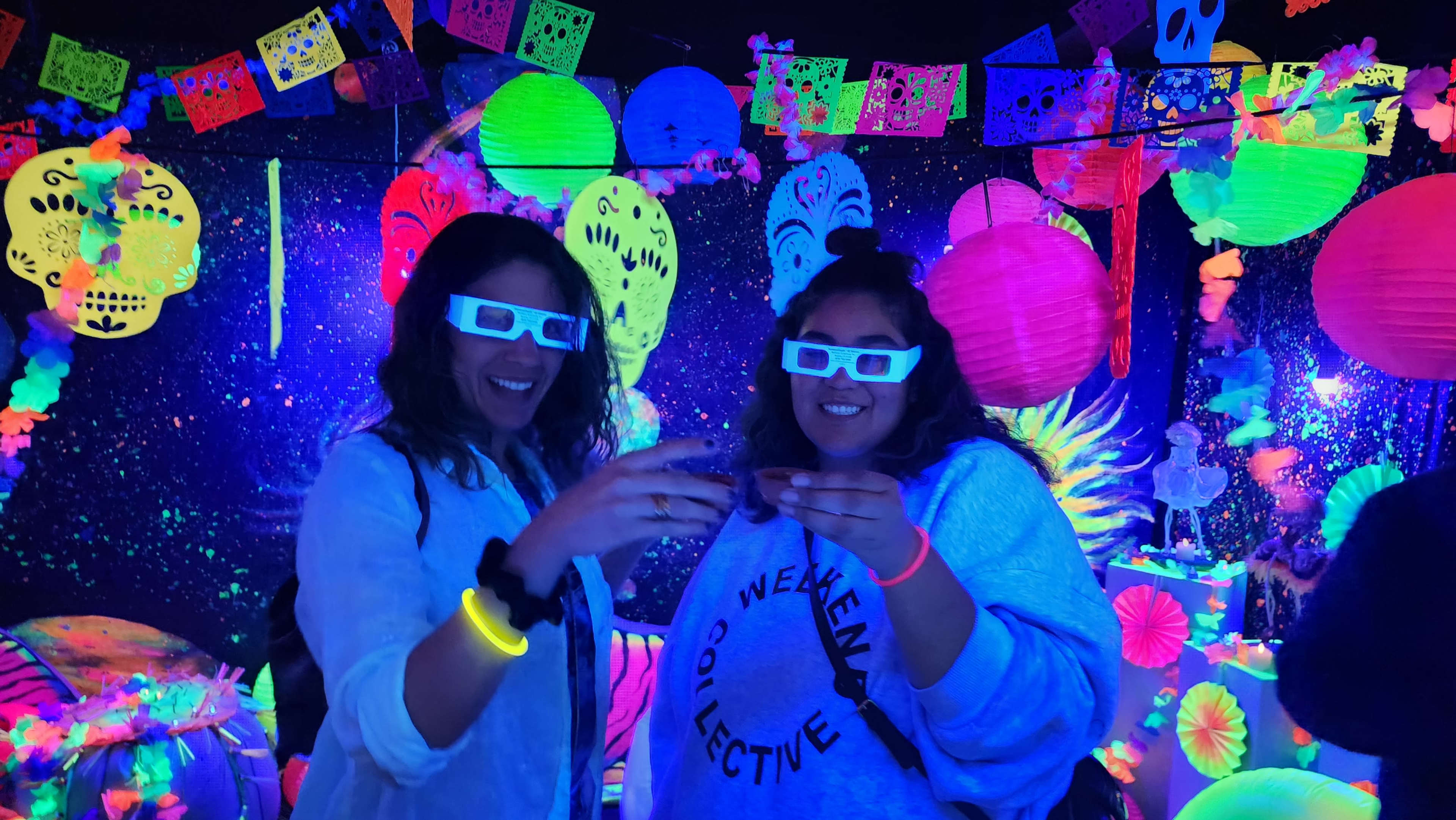 Two women wearing 3D glasses hold drinks in a brightly colored, neon-lit setting decorated with paper lanterns and festive banners.