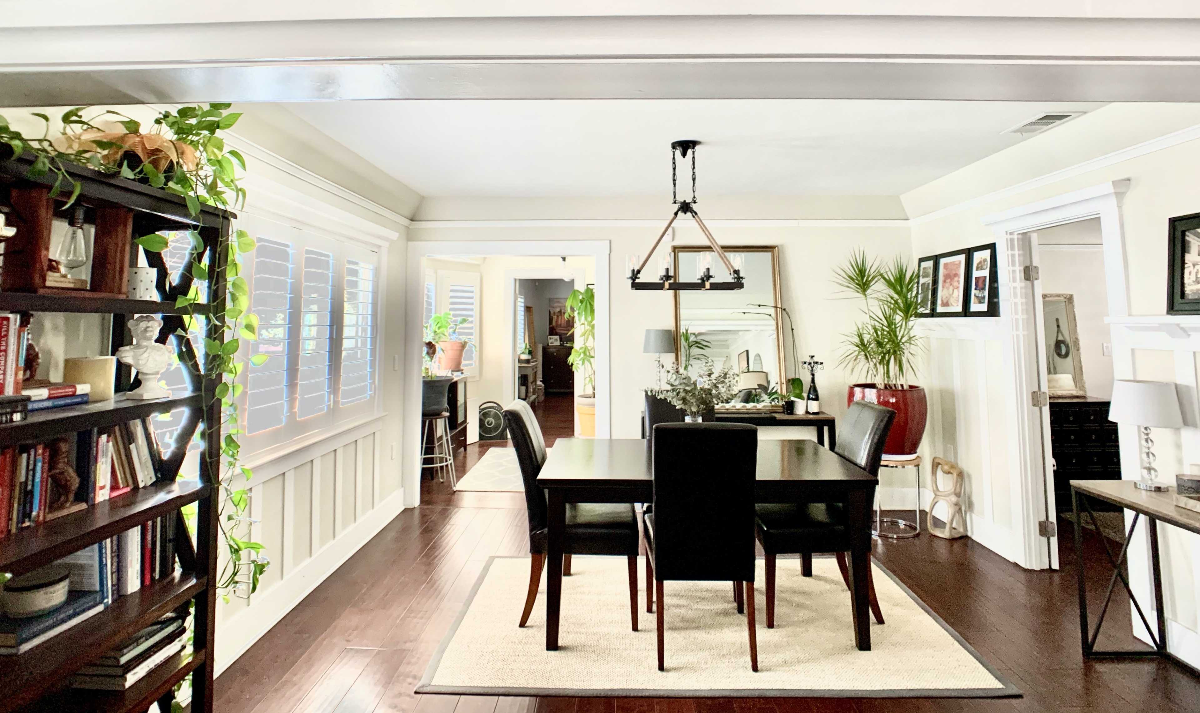 A spacious dining area features a rectangular table surrounded by four black chairs, with shelves on one side displaying books and plants, and doorways leading to other rooms.