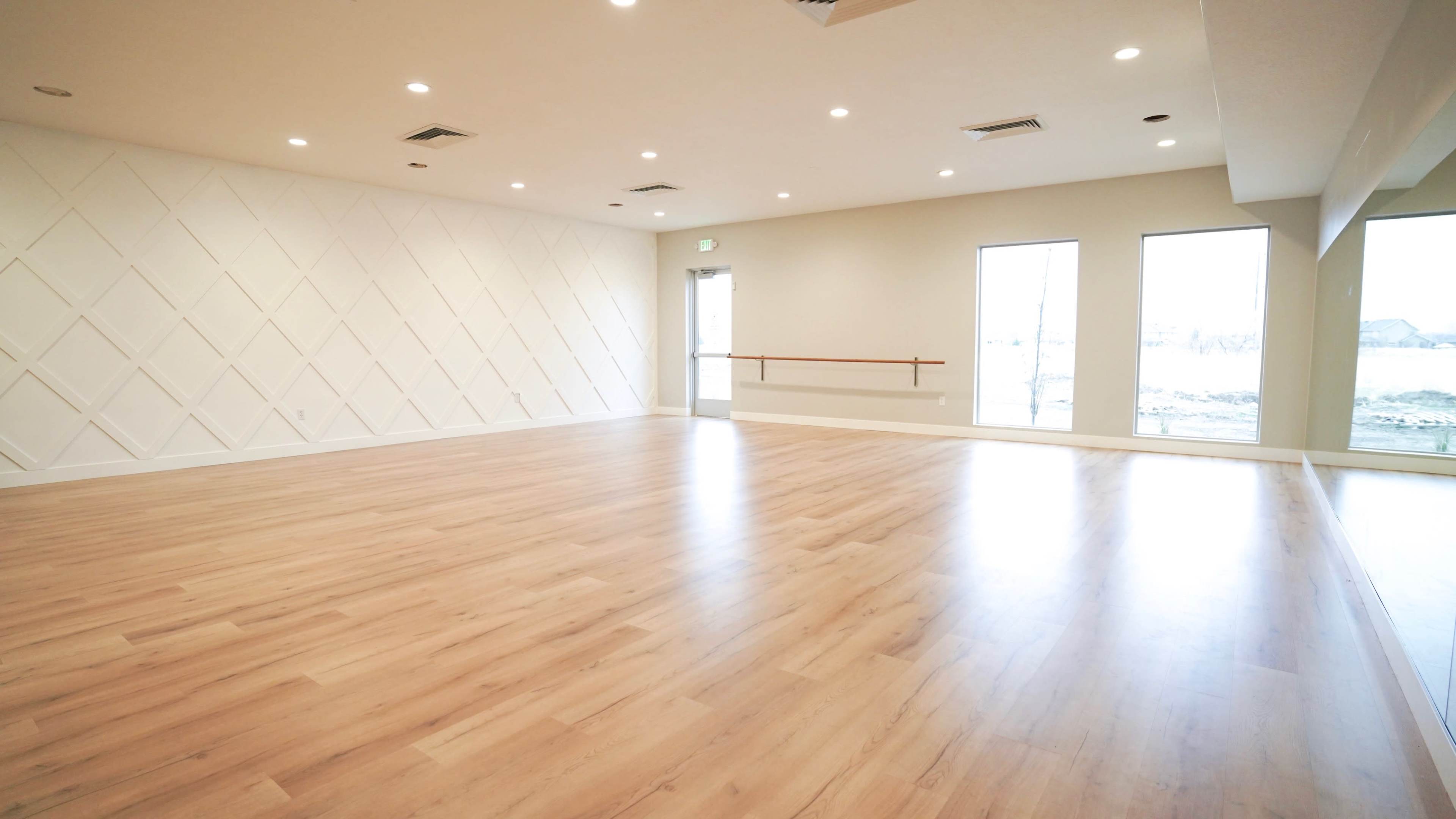 A spacious room with polished wooden flooring, a textured white wall, and large windows allowing natural light to enter.