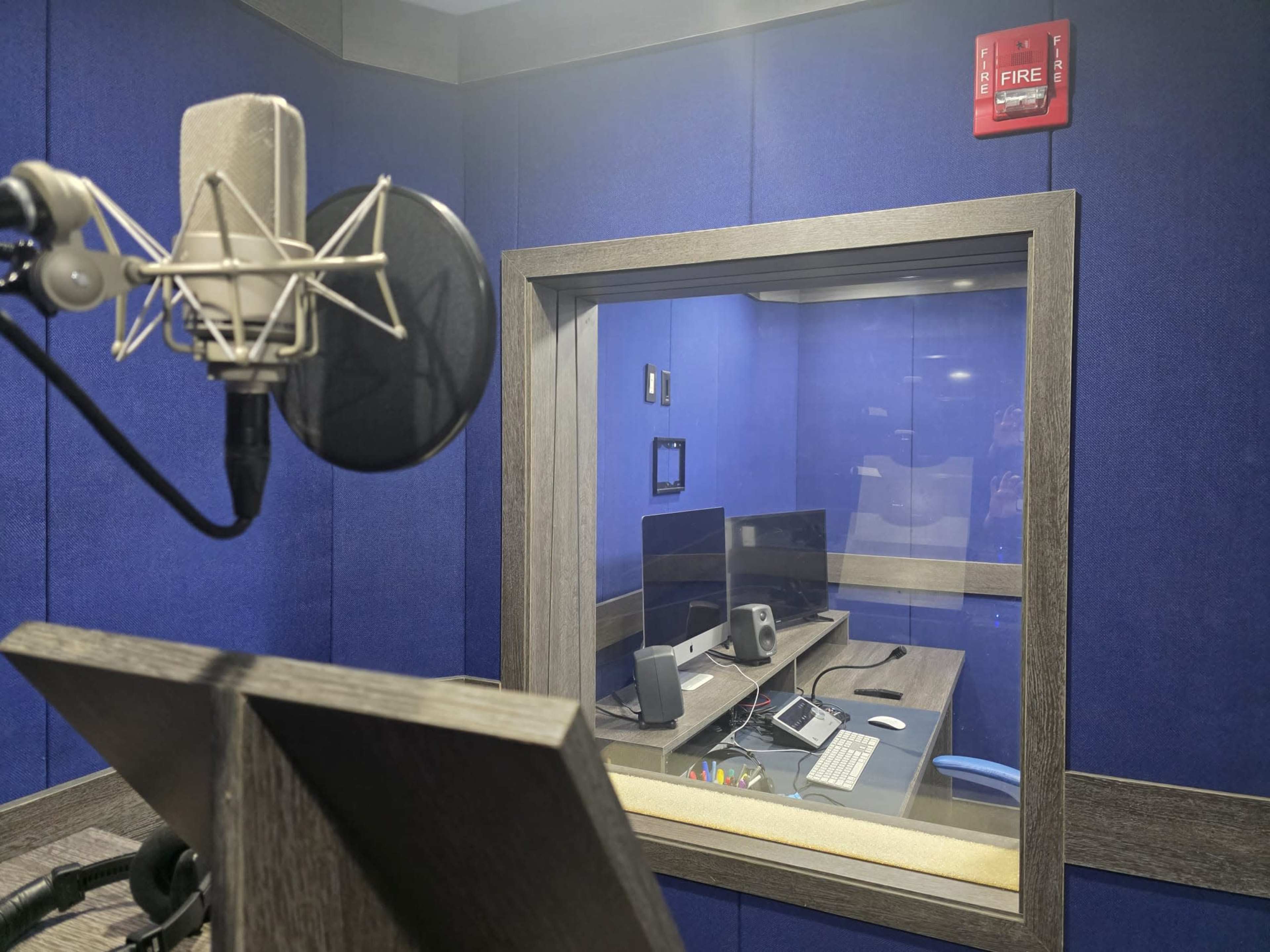 The image shows a sound recording studio with a microphone and a console visible through a glass window.