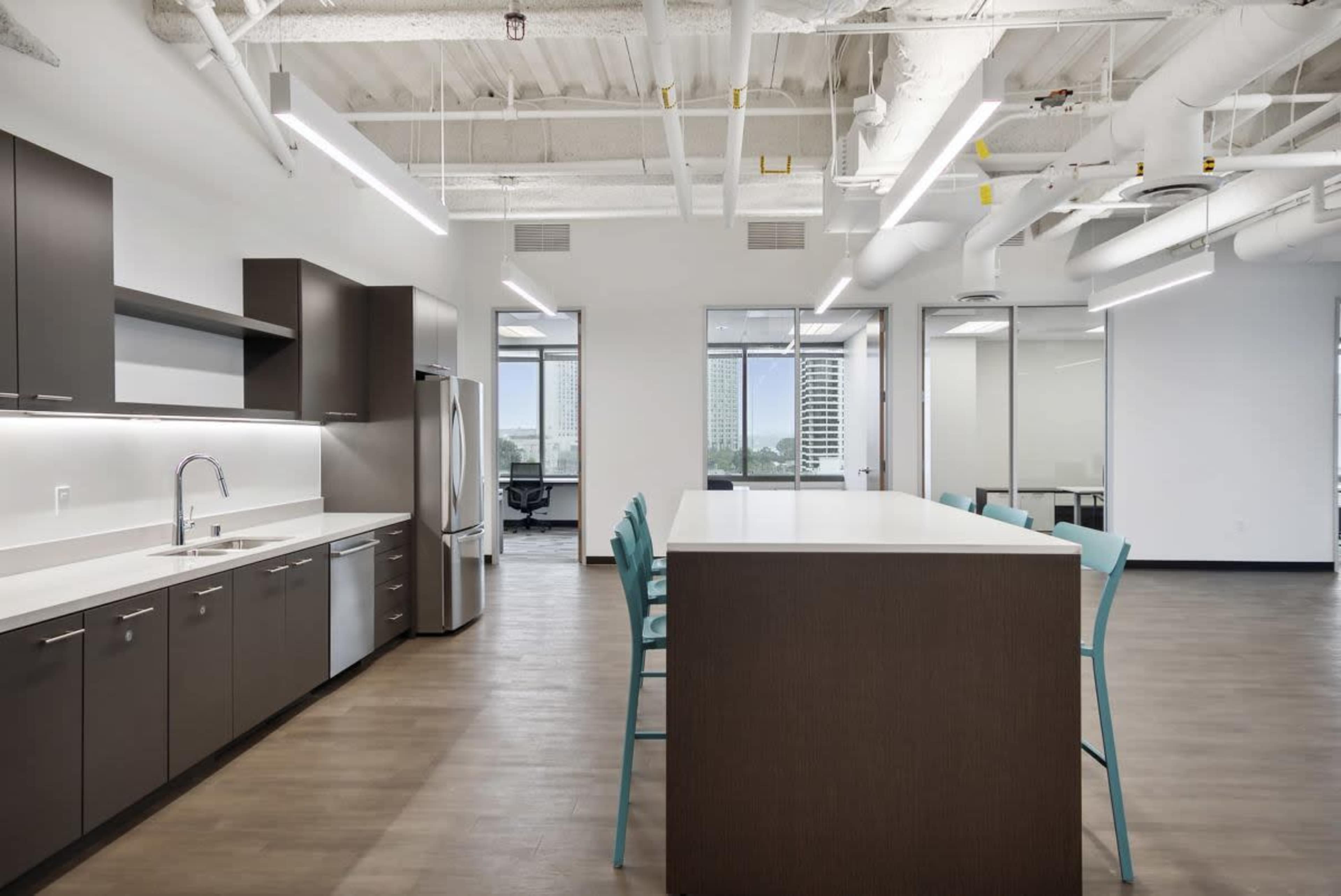A modern kitchen area with a large island, wooden cabinetry, stainless steel appliances, and office spaces visible through glass walls.