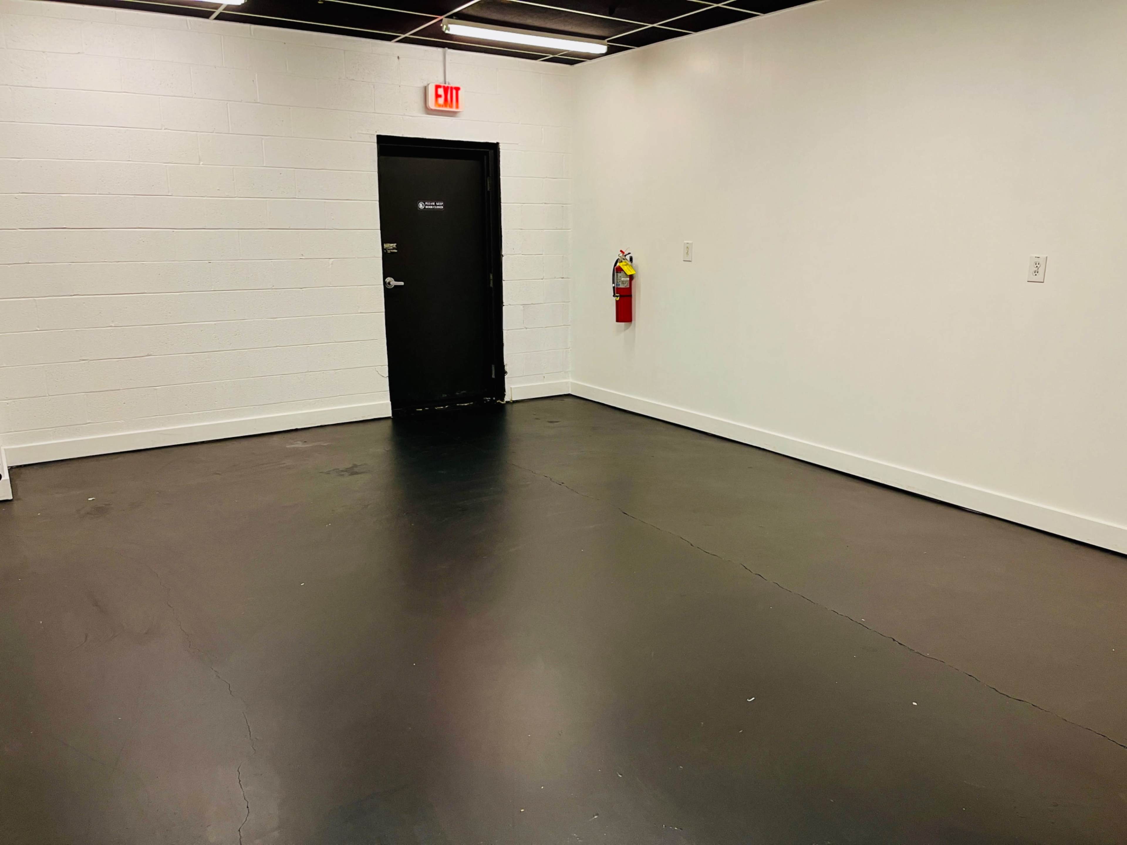 An empty room with black flooring, a white brick wall, a closed black door, and a fire extinguisher mounted on the wall.