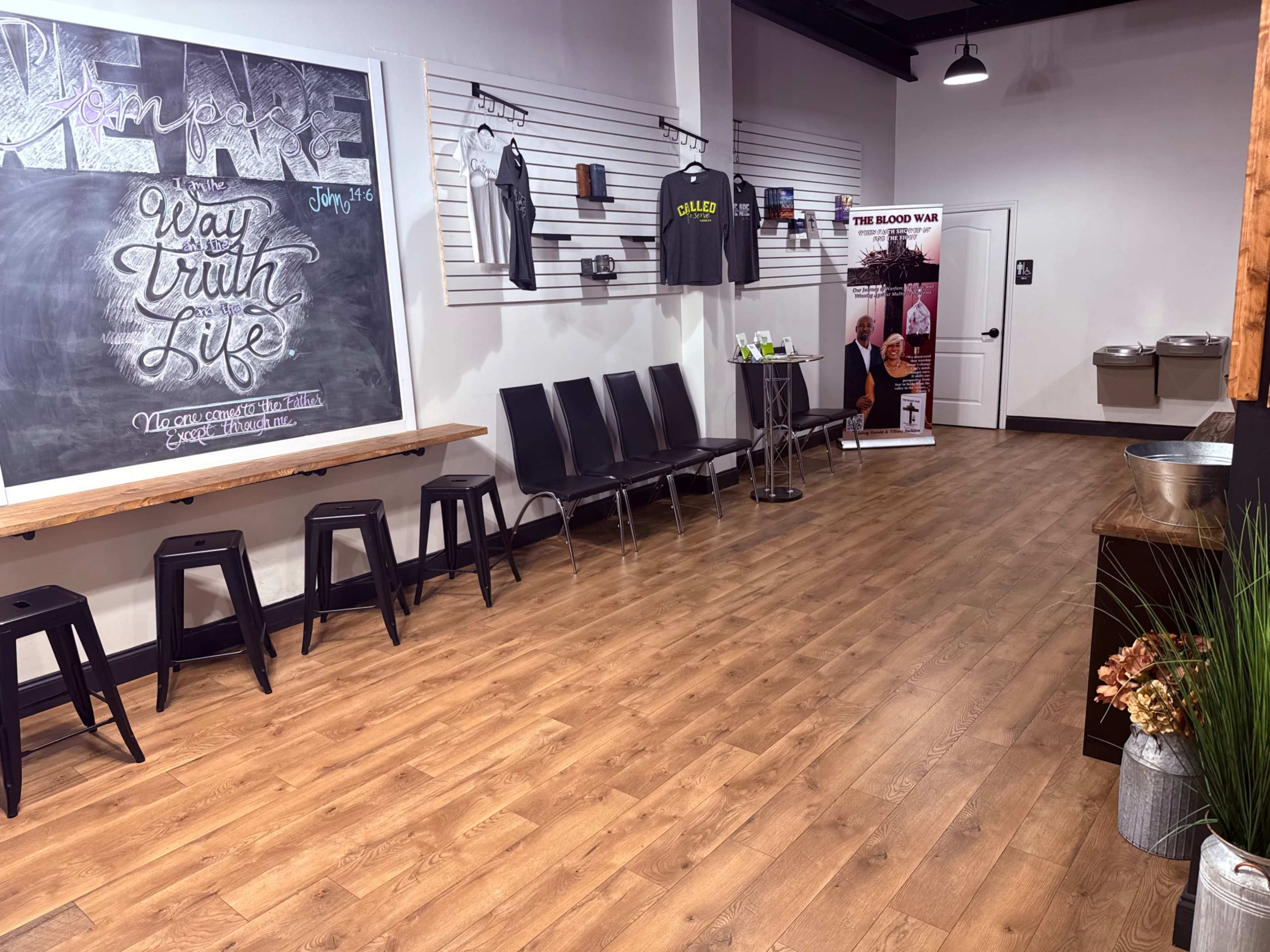 The image shows a spacious interior with wooden flooring, a chalkboard wall displaying text, clothing items hanging on a display, and a row of black chairs beside a refreshment area.