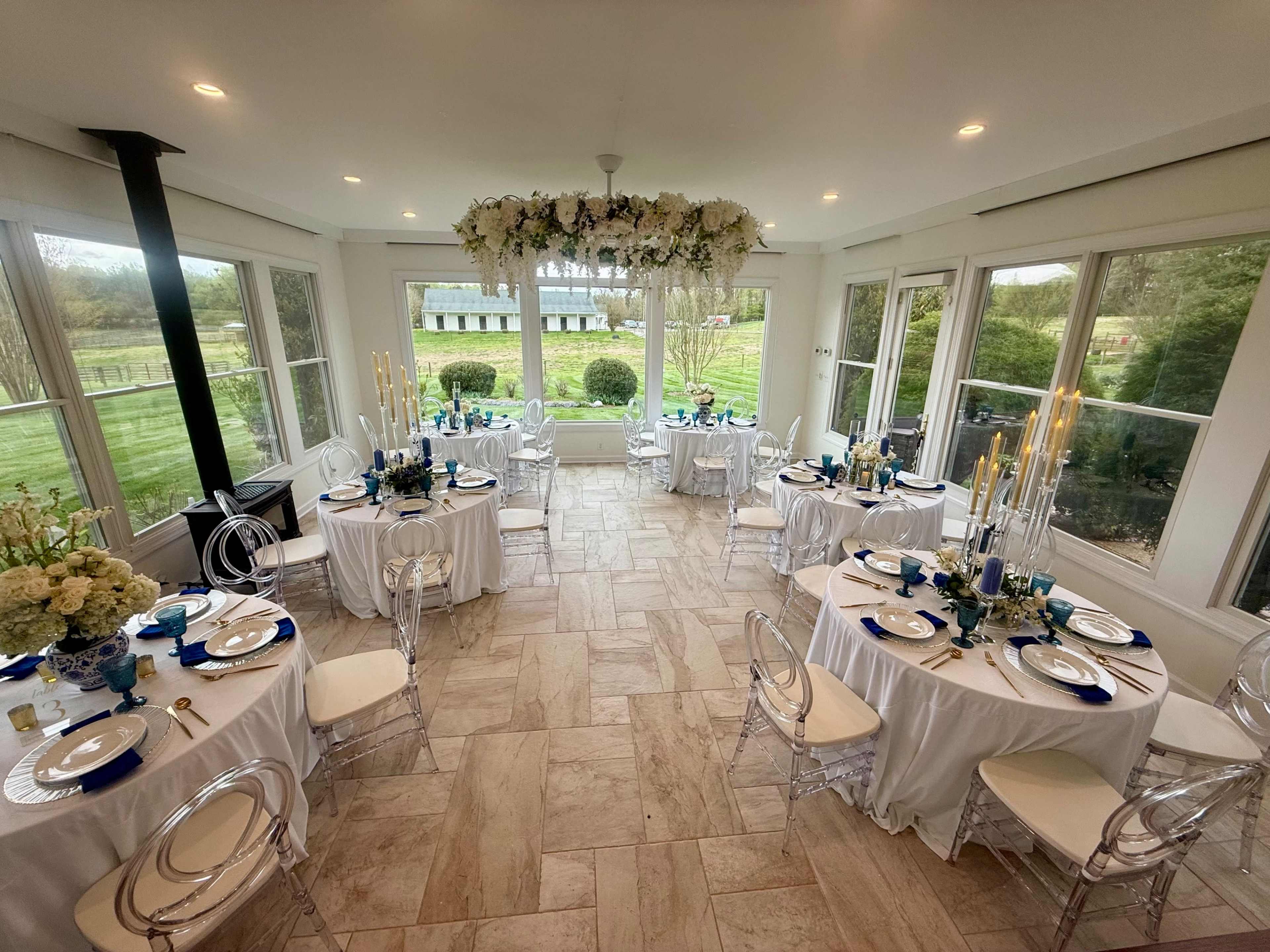The image shows a bright, elegant dining room set for a formal event, featuring round tables with white tablecloths and blue accents, surrounded by large windows overlooking a green landscape.