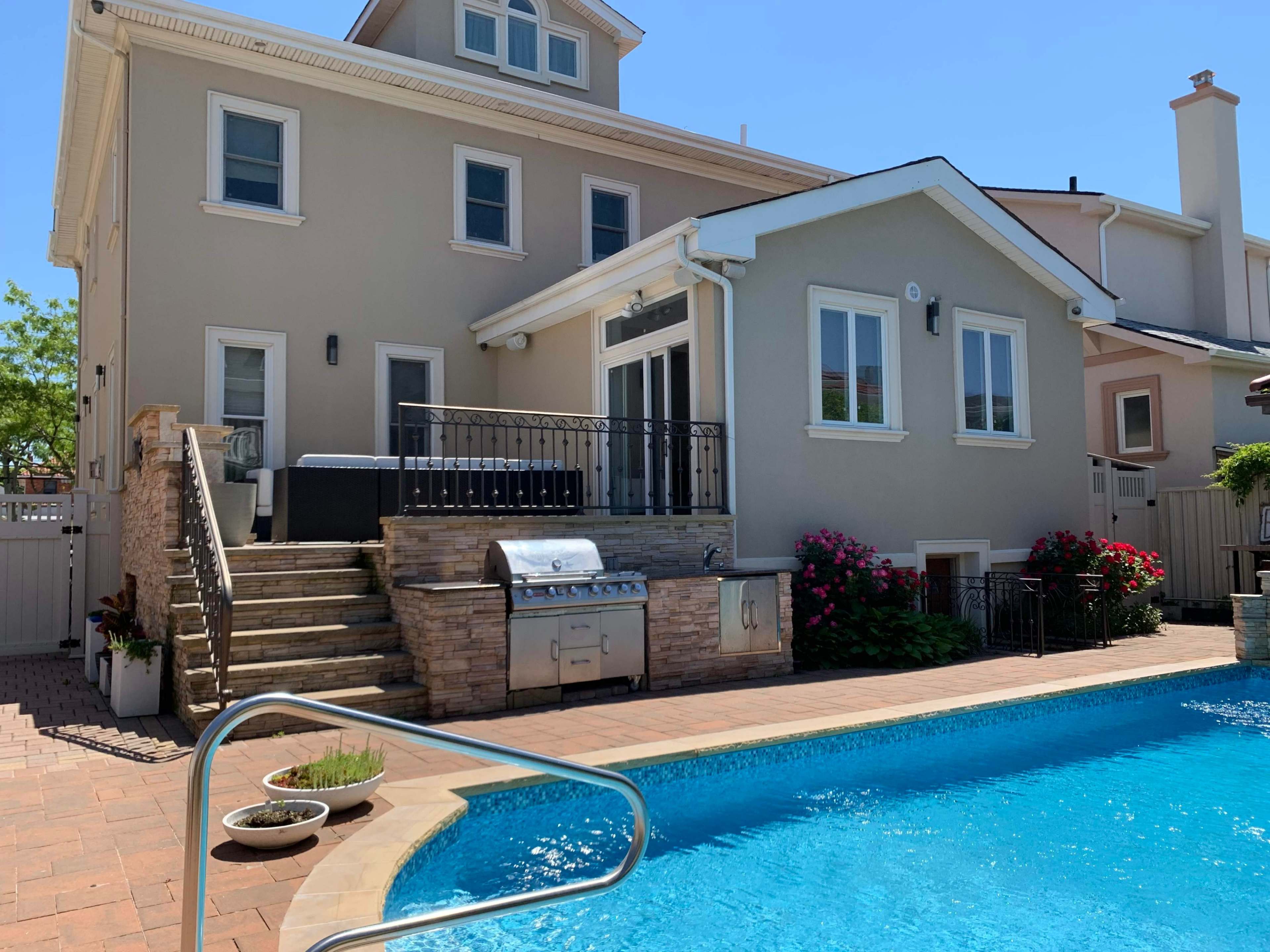 A modern house with a patio and barbecue area overlooks a swimming pool surrounded by landscaped greenery.
