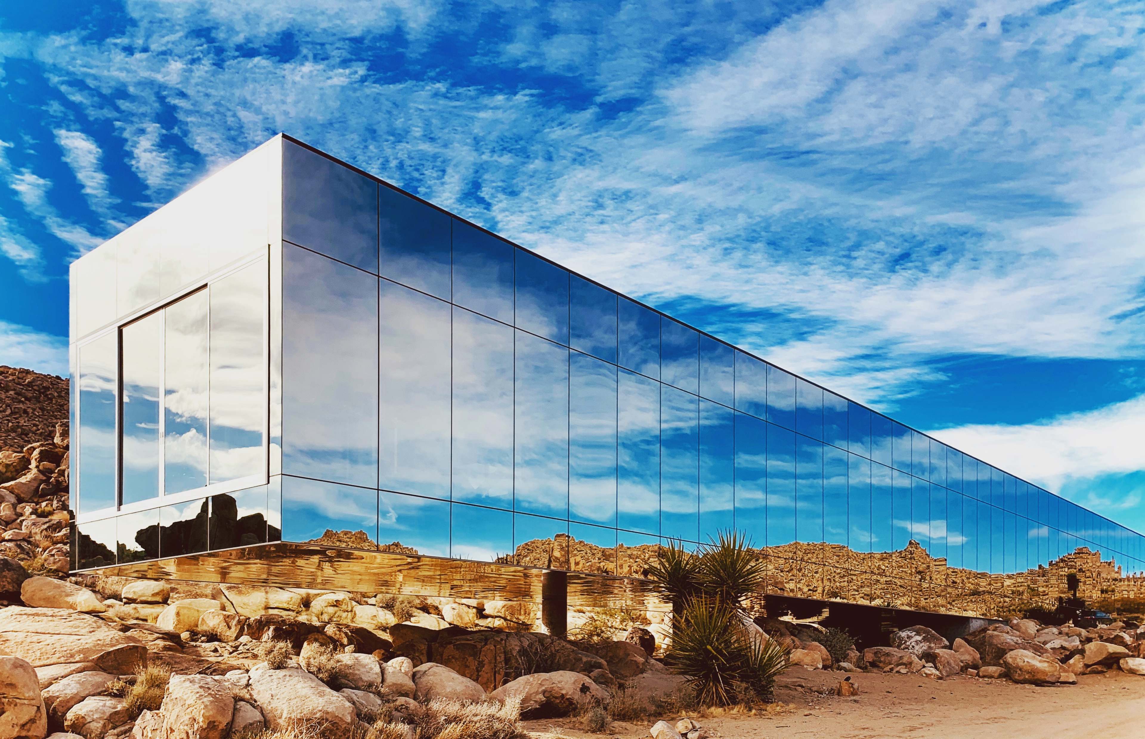 A modern, mirrored building reflects its surrounding rocky landscape and blue sky in a desert setting.