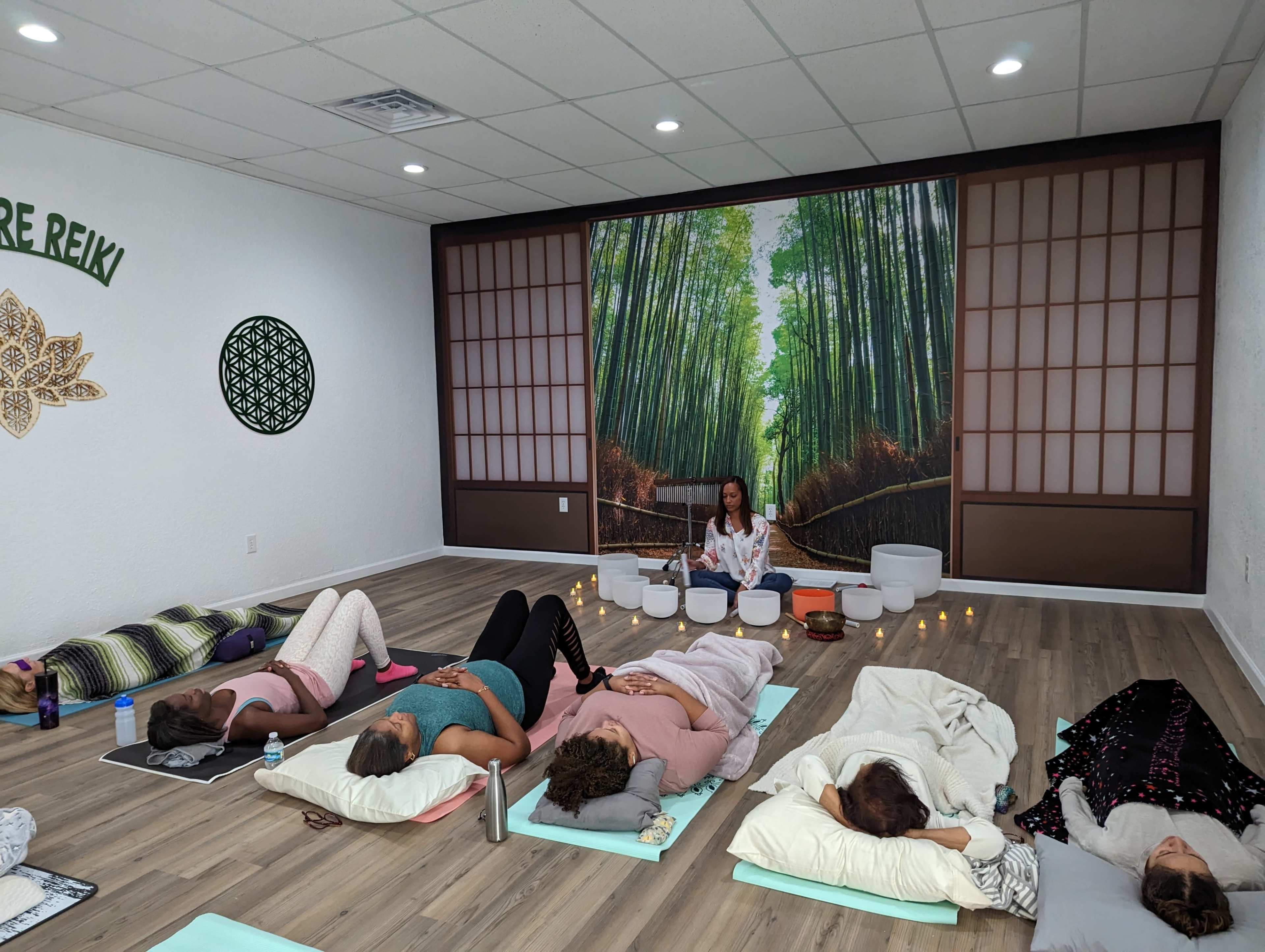 A group of individuals lies on mats in a yoga studio, while an instructor sits at the front with singing bowls in a serene setting adorned with a bamboo forest mural.