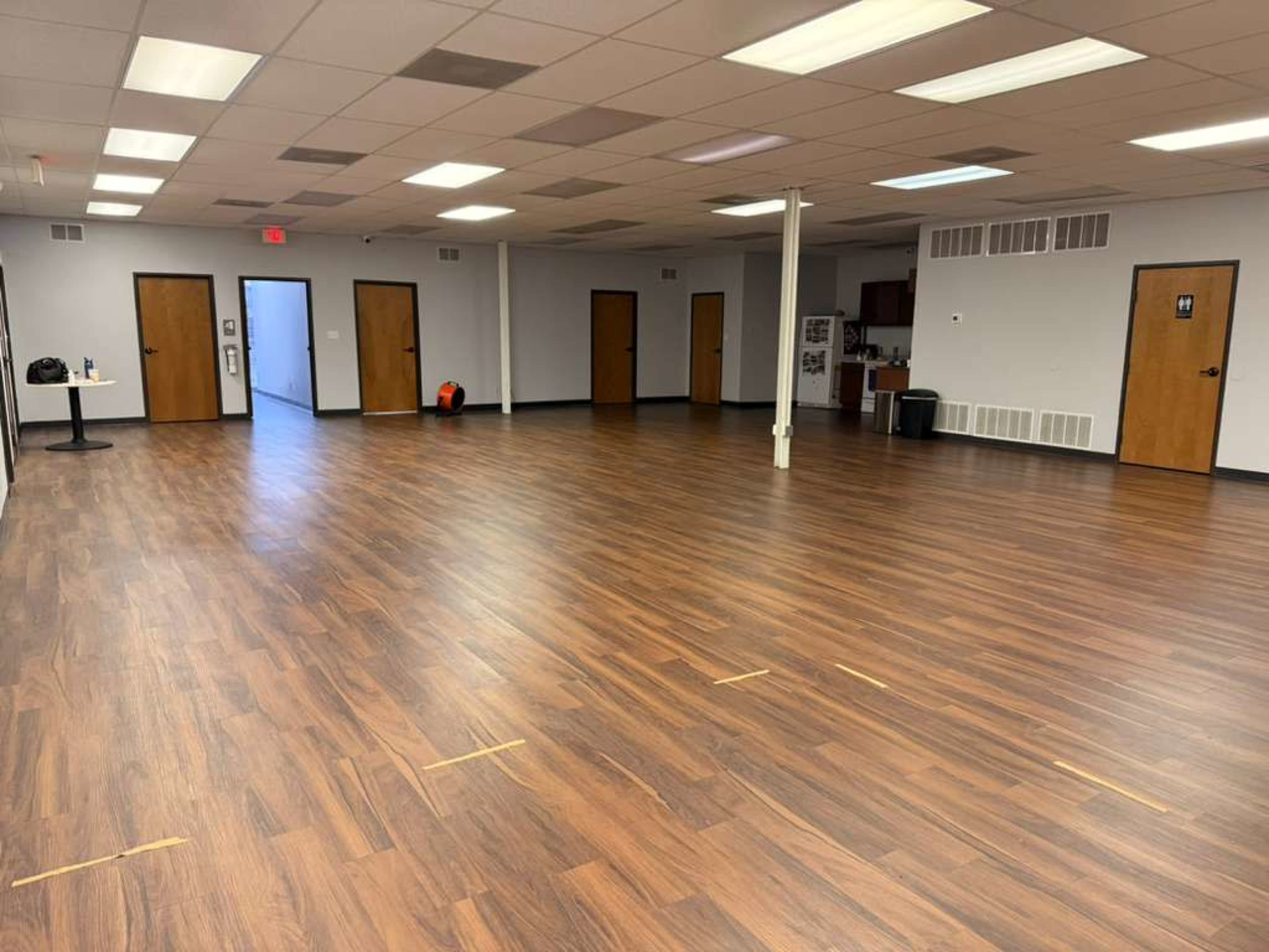 The image shows a spacious empty room with wooden flooring, several closed doors along the walls, and a kitchenette area visible in the background.