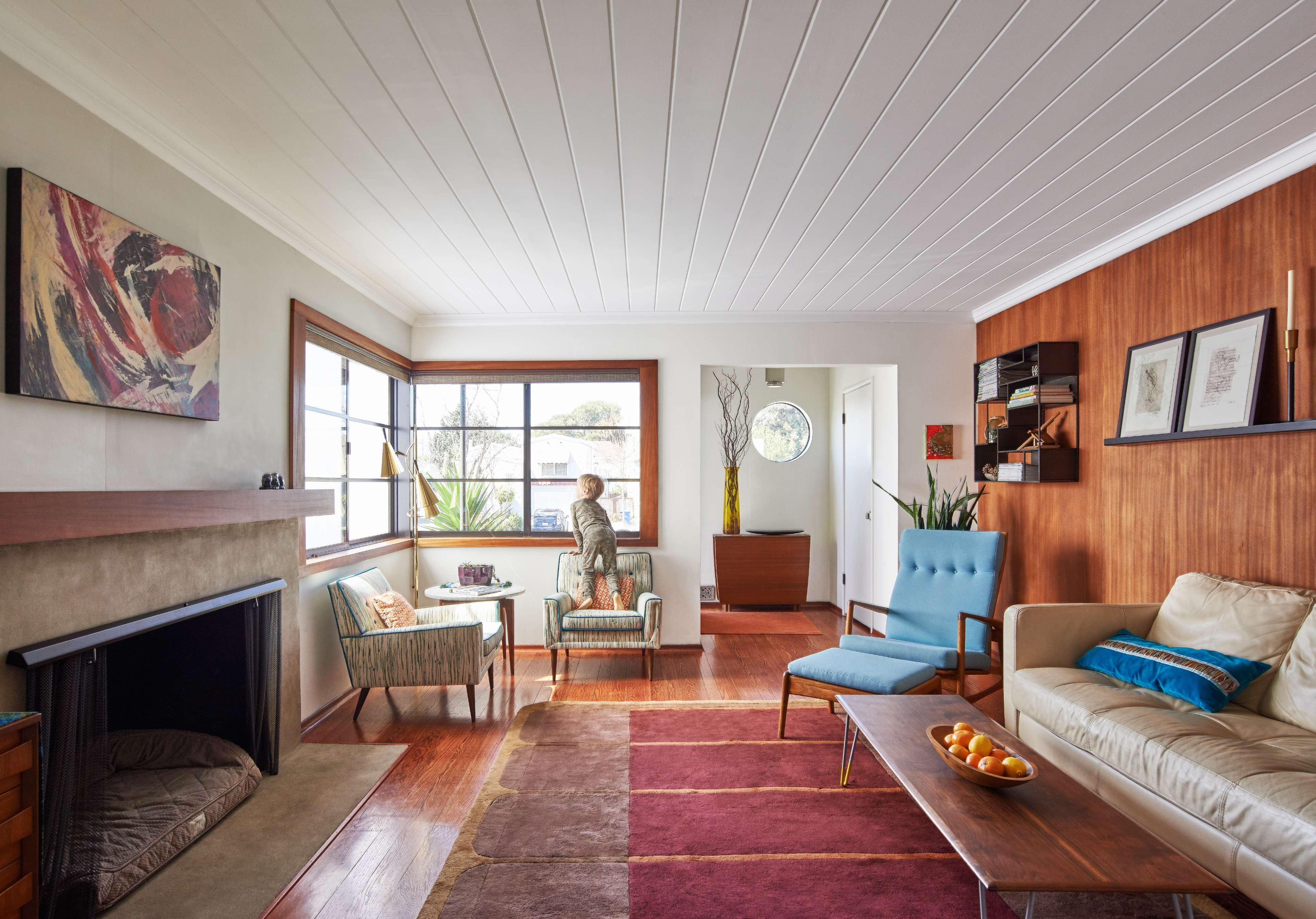 A modern living room with a fireplace, large windows, and a woman sitting by the window, surrounded by minimalist furniture and decor.