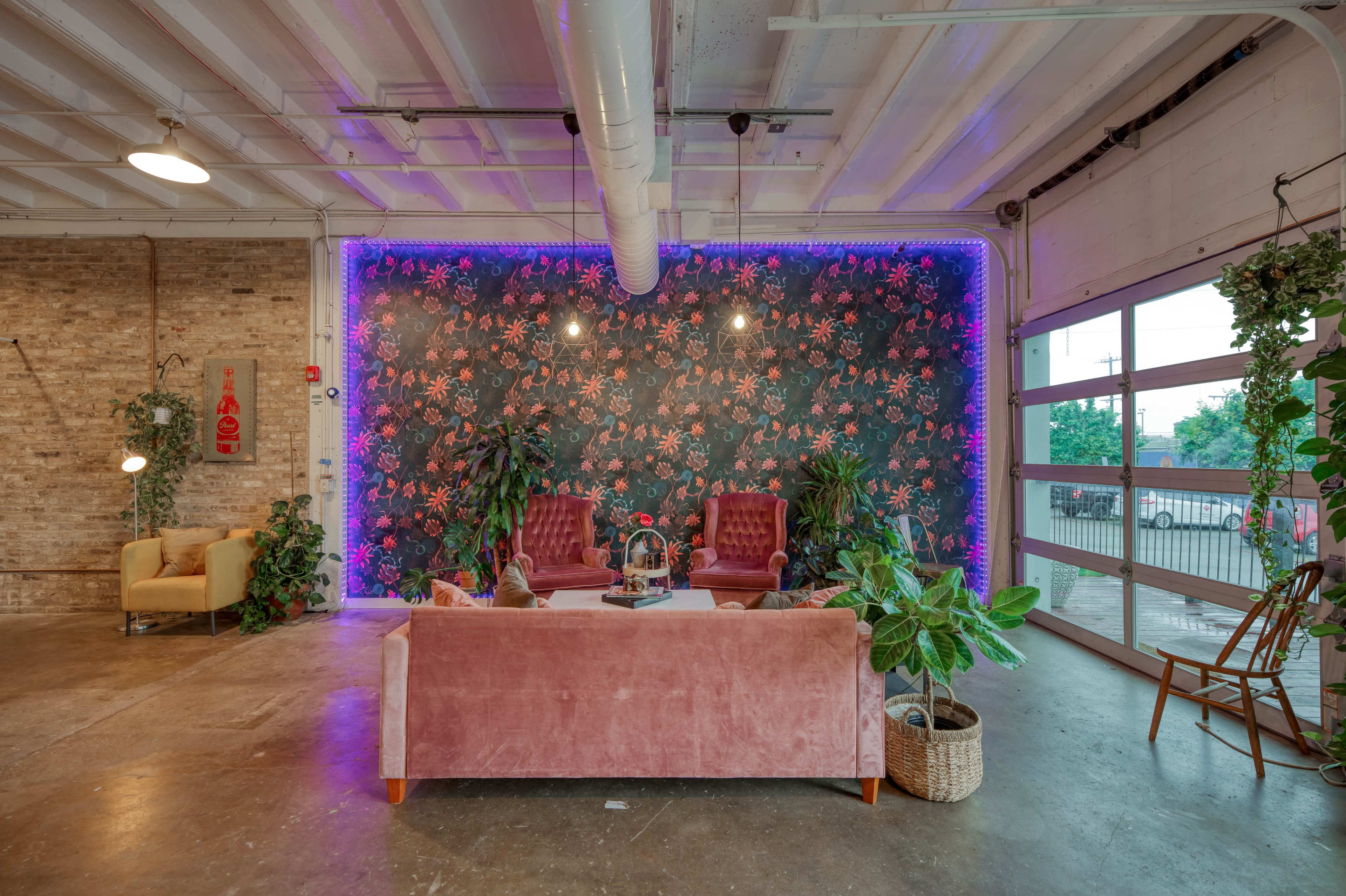A modern interior space with a floral wallpaper backdrop, two red armchairs, a pink sofa, and various plants, illuminated by purple lighting.